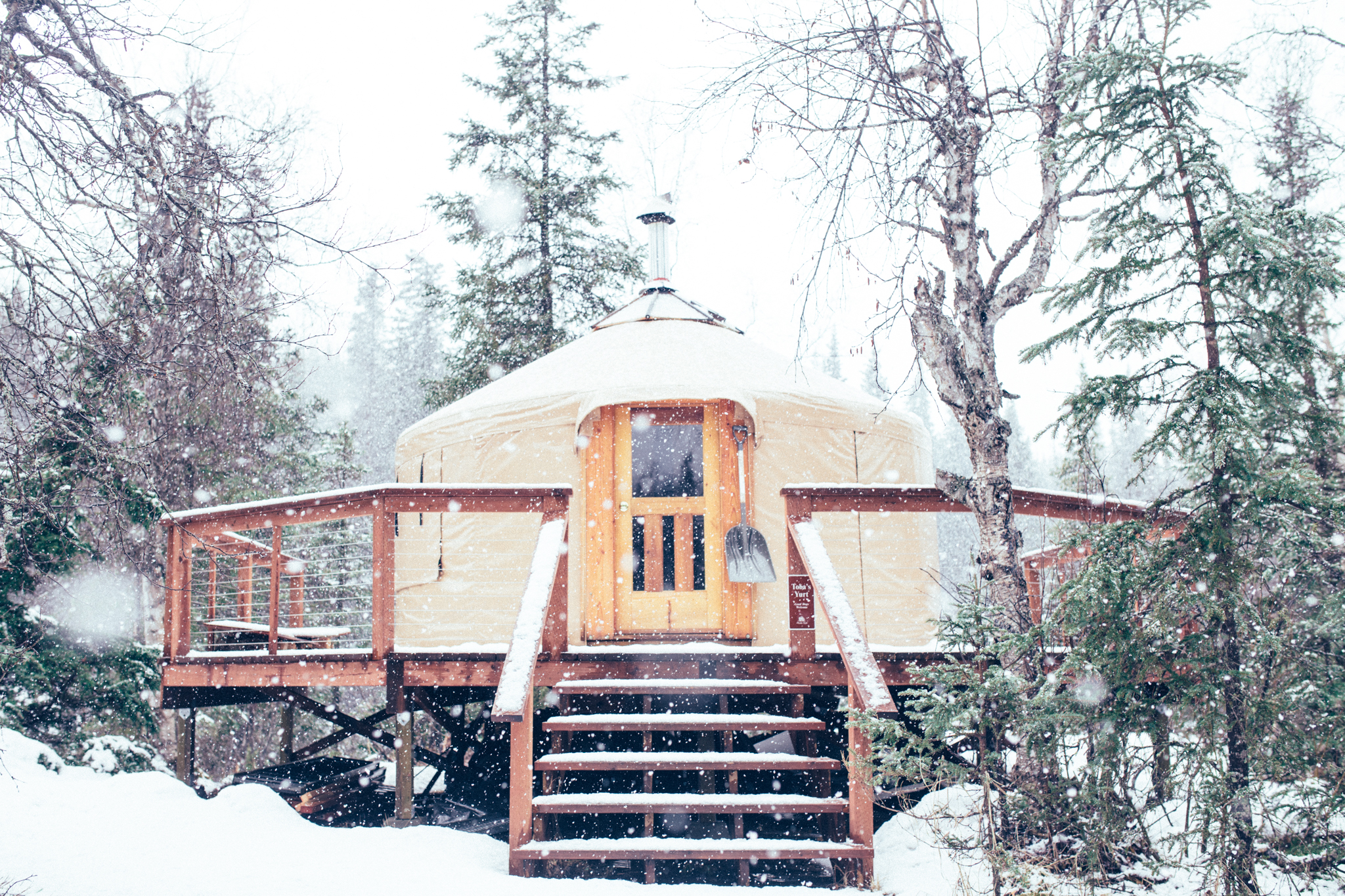 A snowy yurt surrounded by a porch and railings with a central staircase sits in the woods. 
