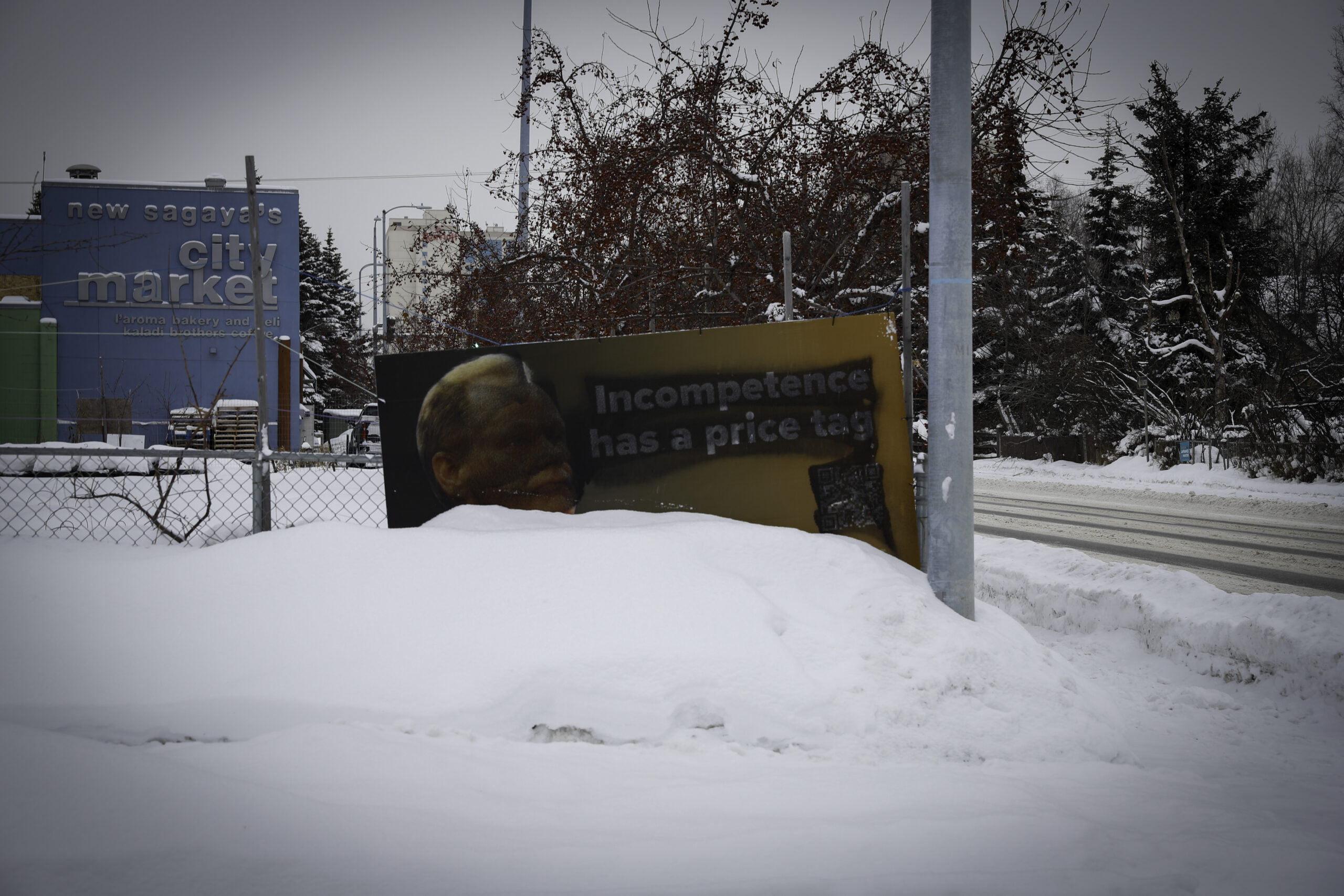A sign that says, "Incompetence has a price tag" is spray painted. It is standing half way buried in snow near a light pole along a wired fence.
