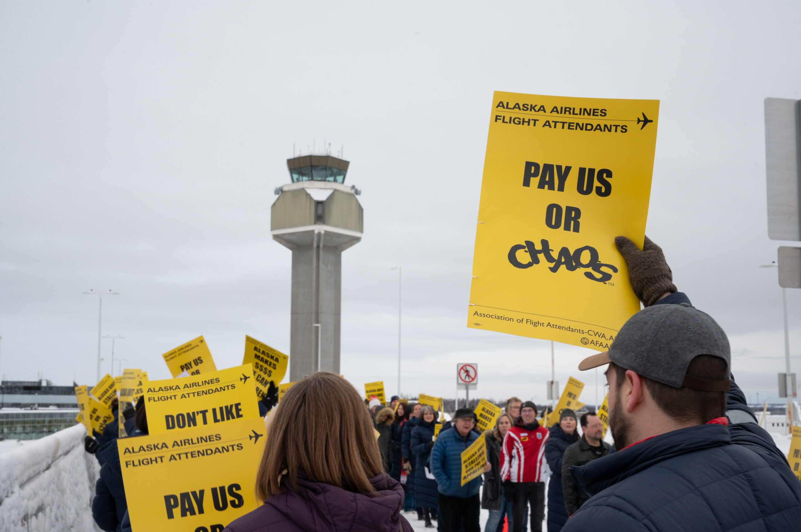 Flight attendants hold signs in a picket line outdoors, with an airport control tower in the background