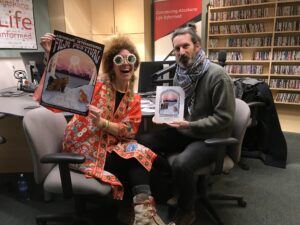 A woman and man pose with posters and a radio studio. 