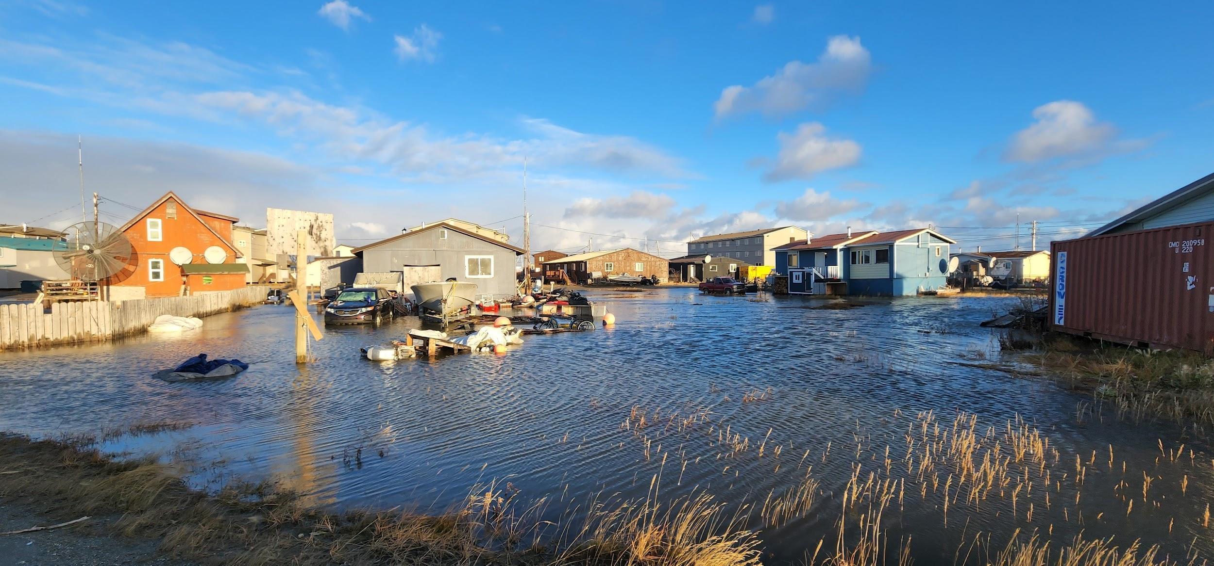 On a sunny day, homes stand in a few feet of flooded, still water.