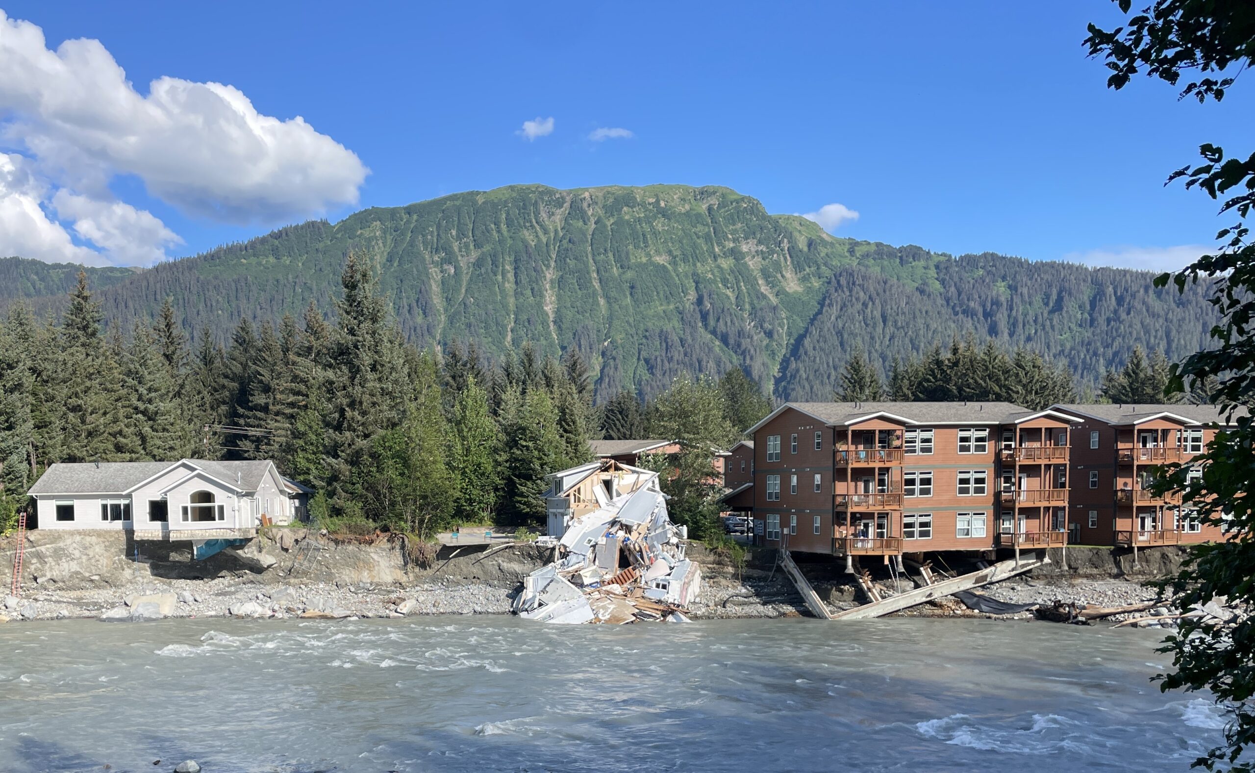 Three residential buildings, including one entirely destroyed home, perch over an eroded riverbank.