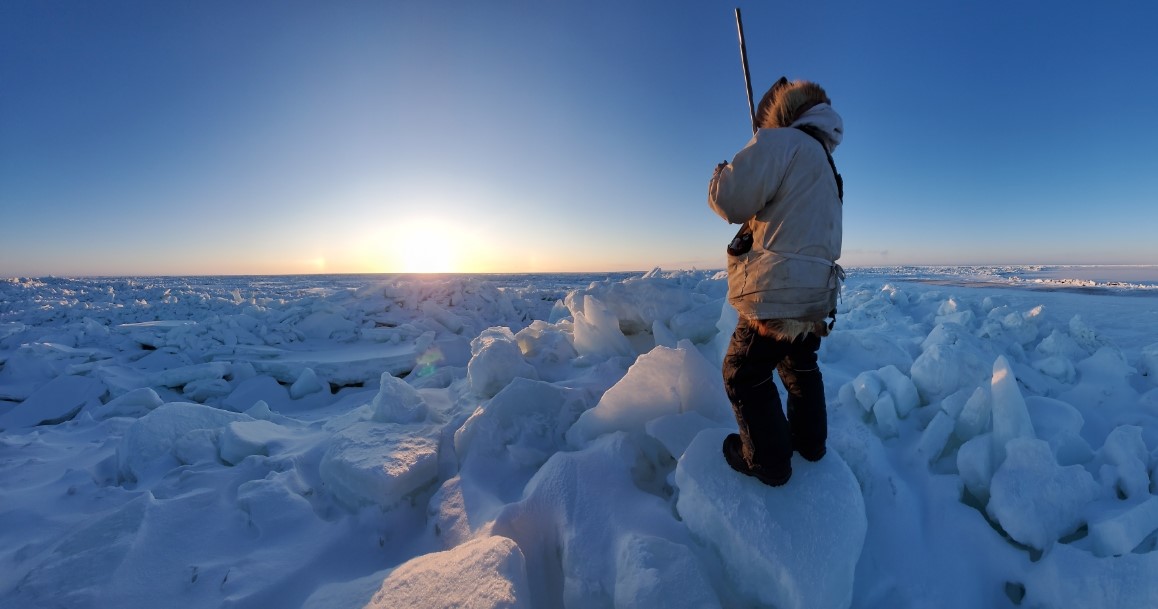 A man in a parka stands on an ice field looking toward the sun low in the sky.