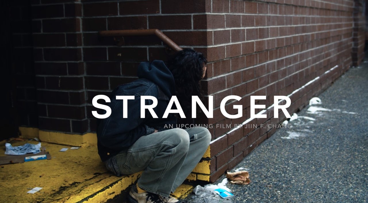 A man sits under a stoop with the word STRANGER. 