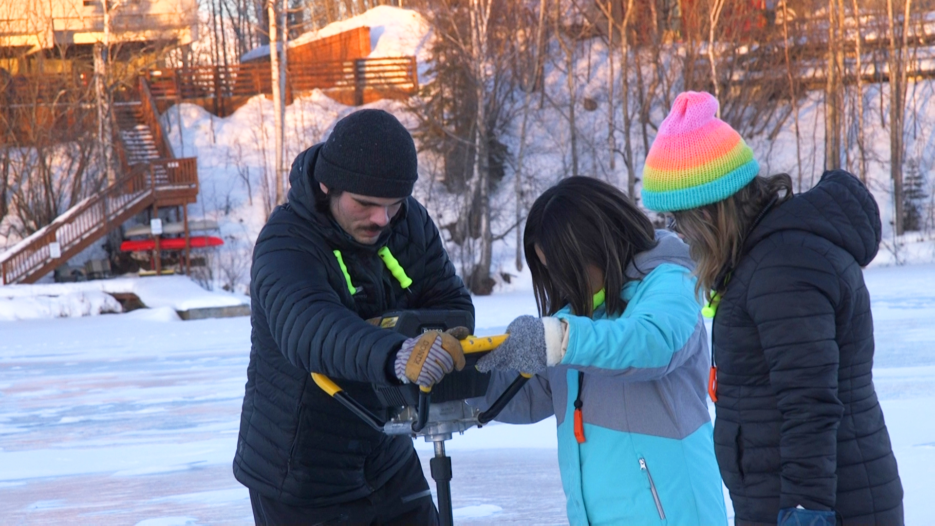 An adult helps two teenage students use an ice auger on Wasilla Lake.