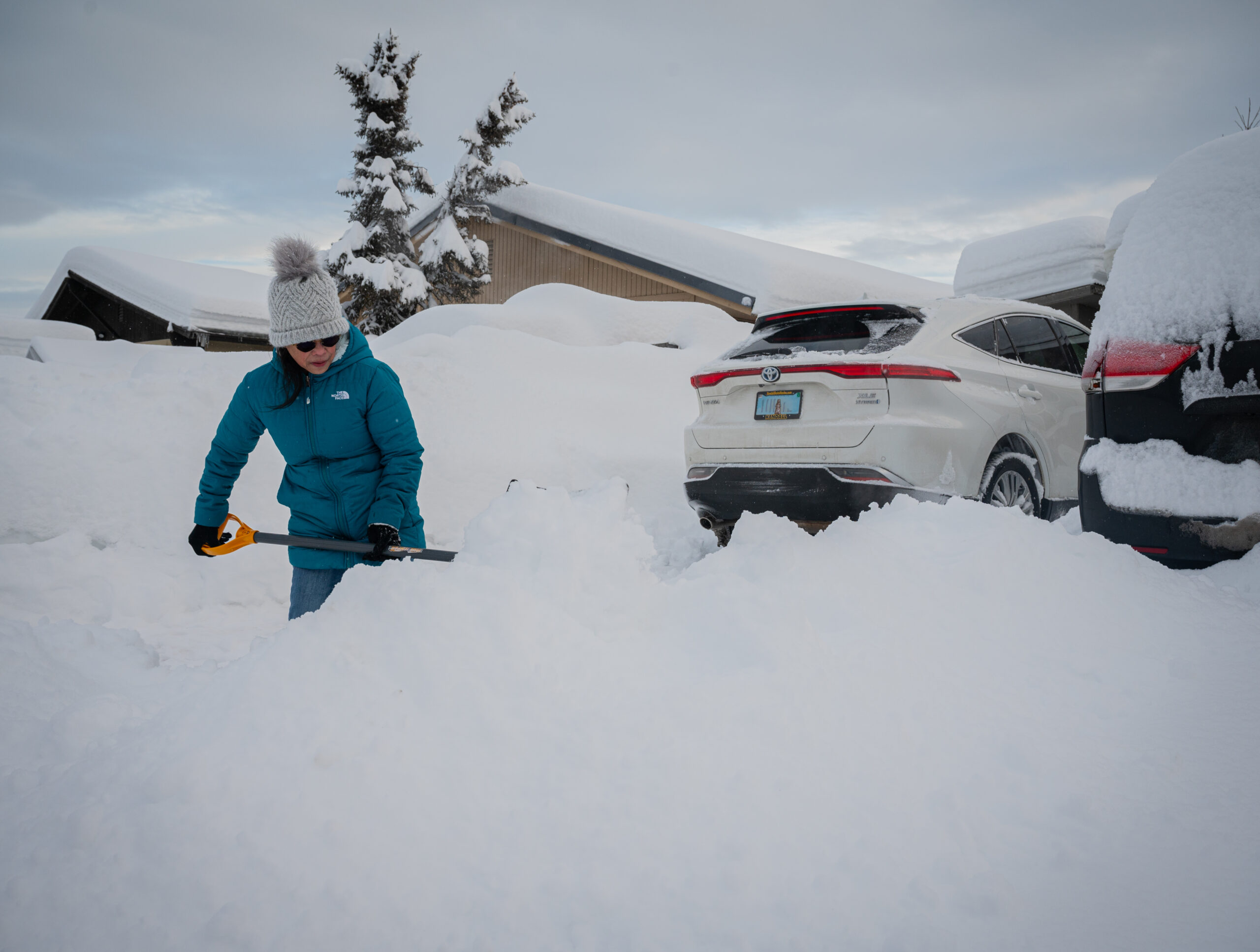 A woman wearing a beanie and blue coat shovels her driveway.