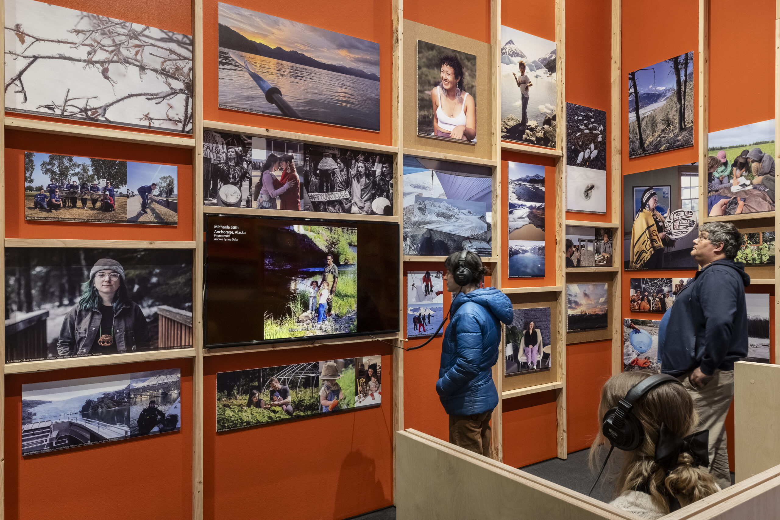 Museum-goers and listen to audio and look at a collection of photos on a red wall.