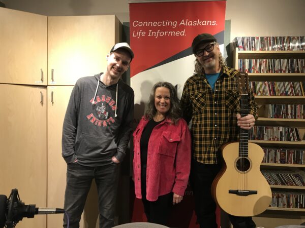 A woman stands between two men, the one on the right is holding a guitar and they are standing in a radio studio.