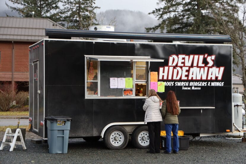 Two people in jackets stand at the window of a food truck that reads "Devil's Hideaway"