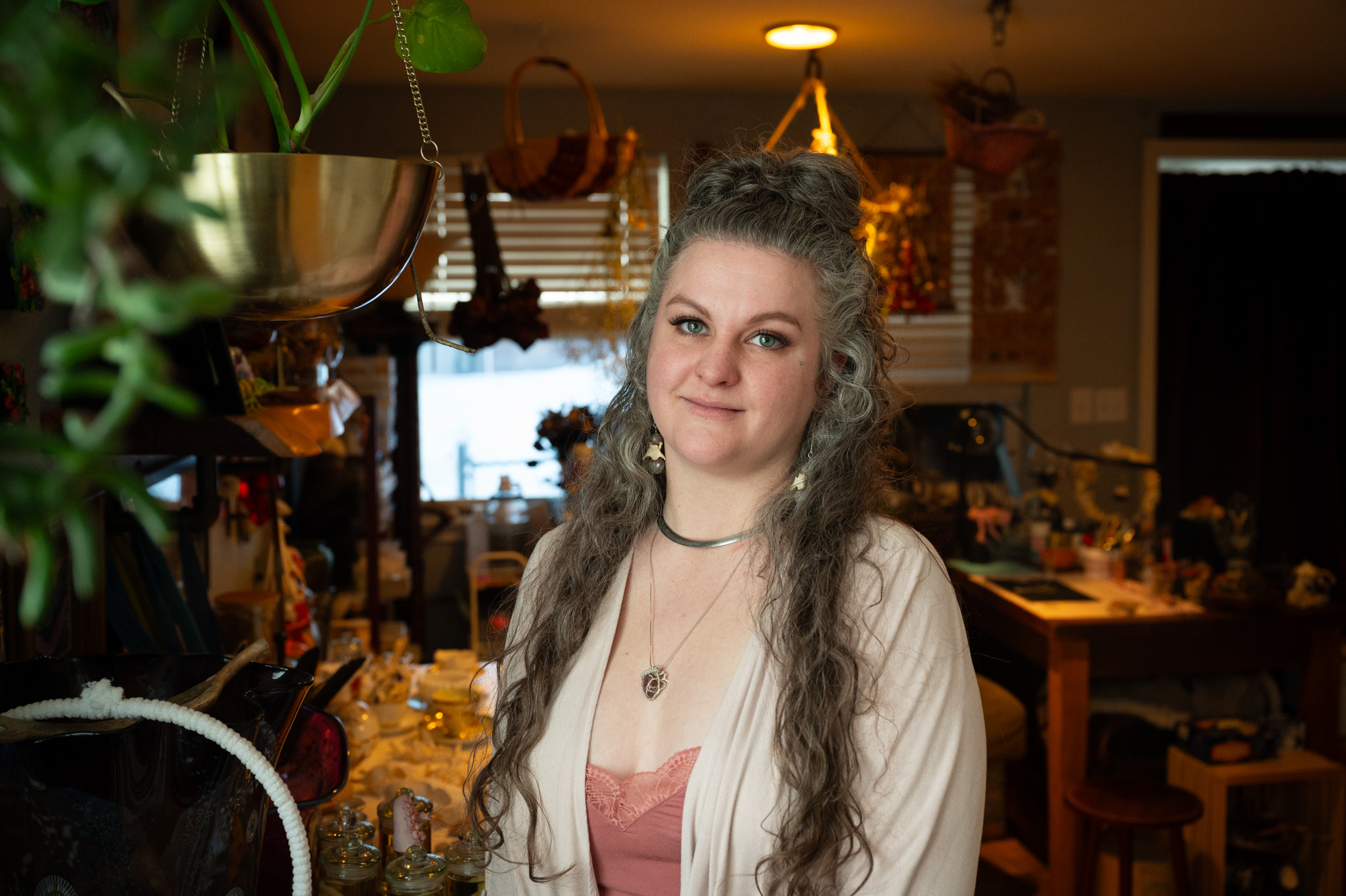 Woman with long hair stands in a room cluttered with plants and others things