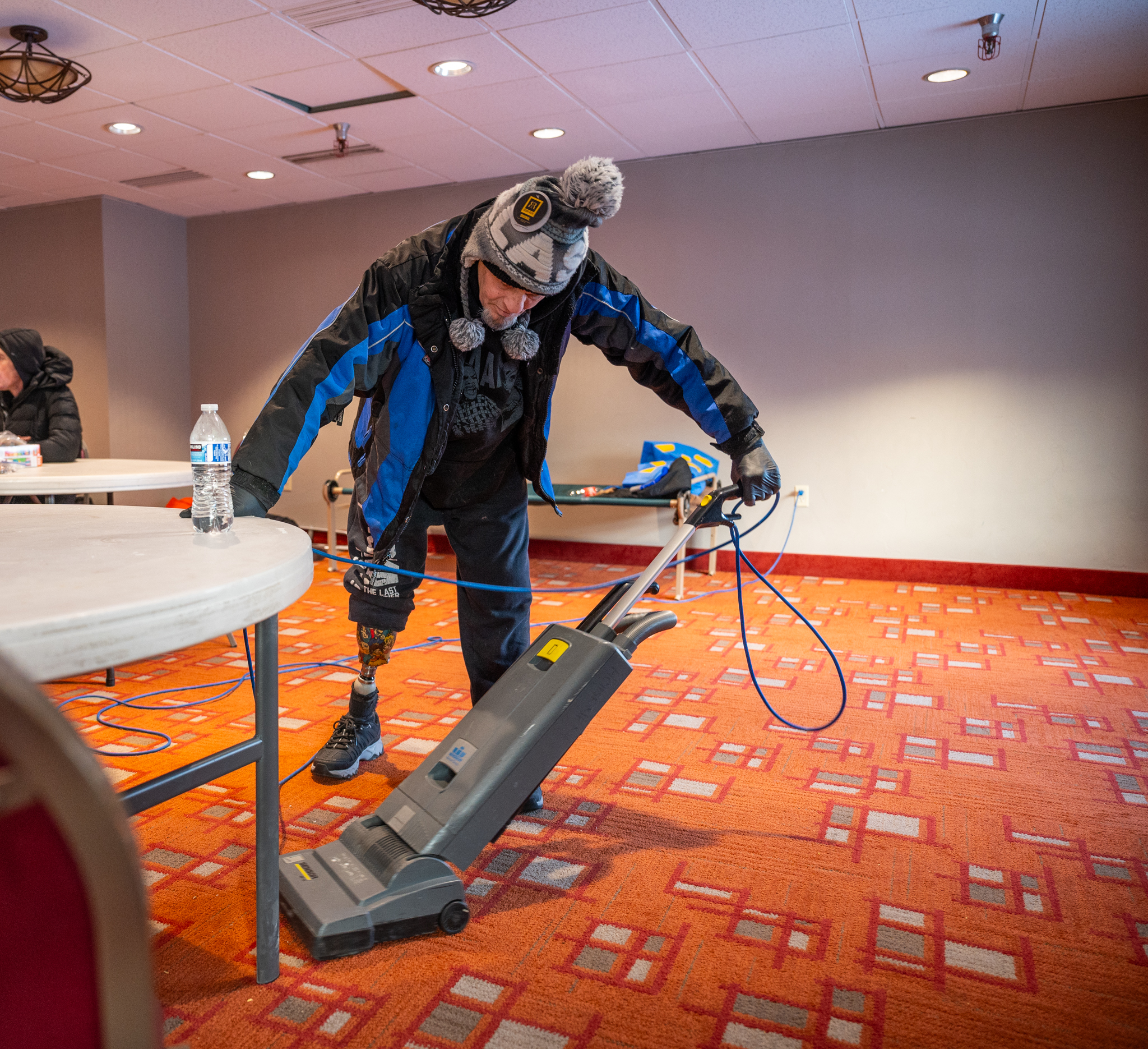 A man with a blue winter coat on vacuums the floor