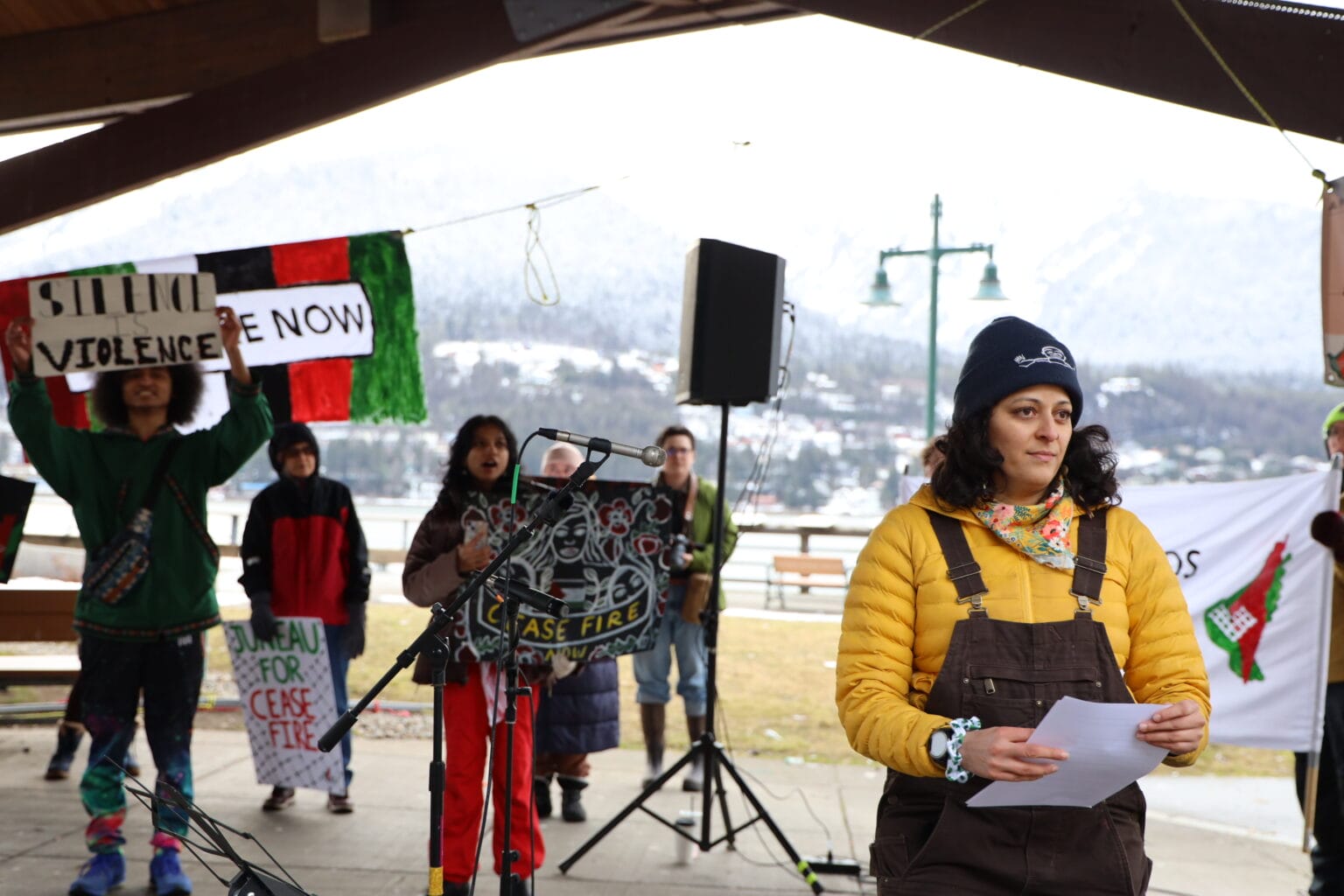 a woman holds a paper in front of a group that has signs that say Juneau for Cease Fire