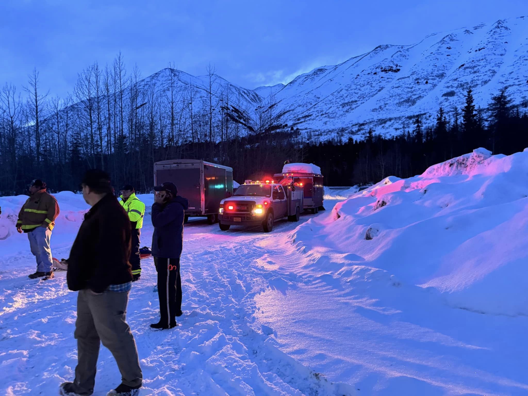Emergency first responders stand in a snowy pullout next to emergency vehicles at dusk. Snow covered mountains in the background.