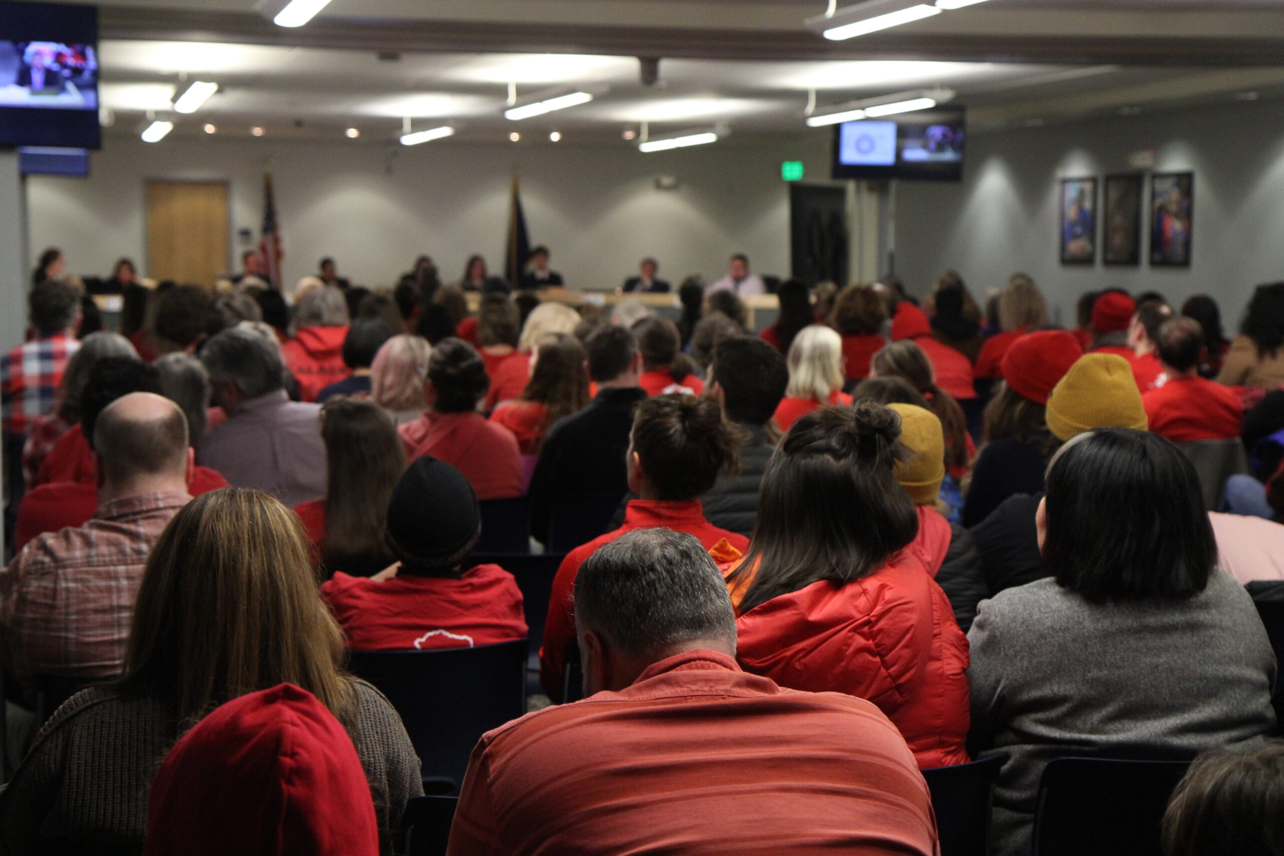 People dressed in red watch a school board meeting.