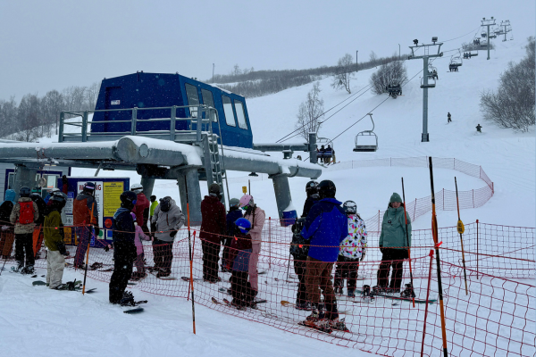 People on skis and snowboards line up in front of a ski lift and mountain.
