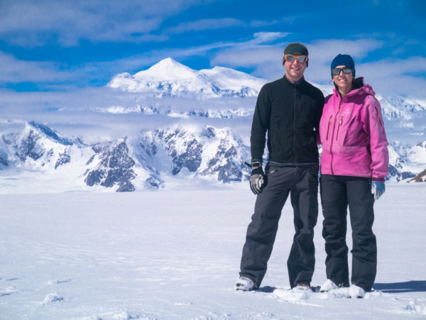 A man and a woman stand in the snow in front of a large mountain. 
