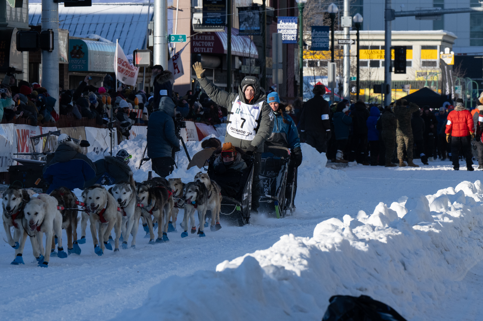 A man in black coat waves to fans on a dog sled.