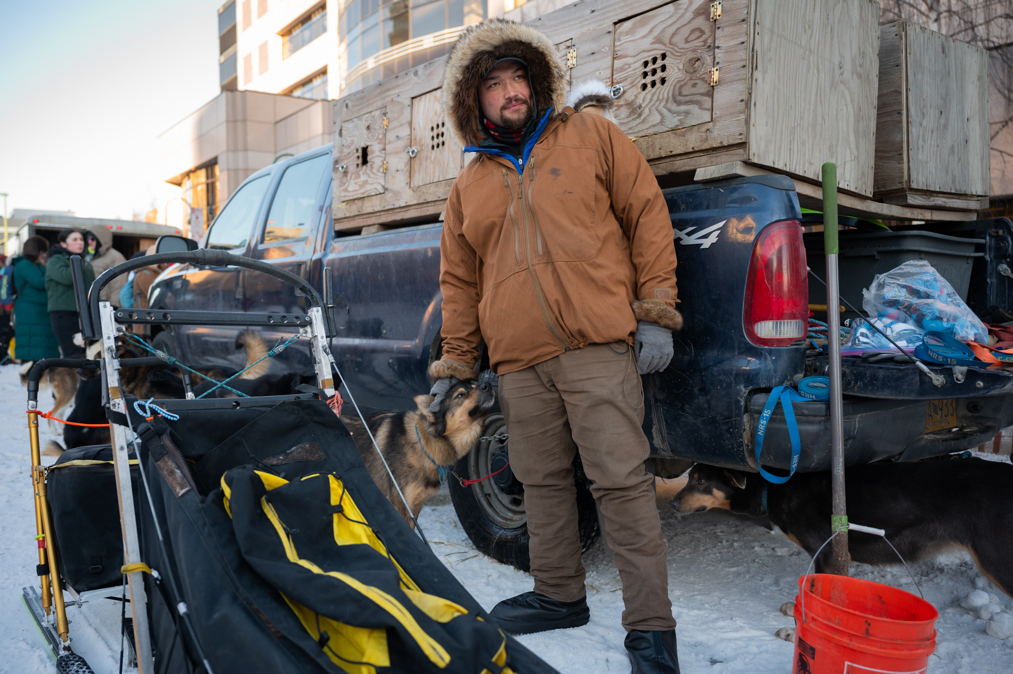 A man in a brown winter jacket pets a sled dog