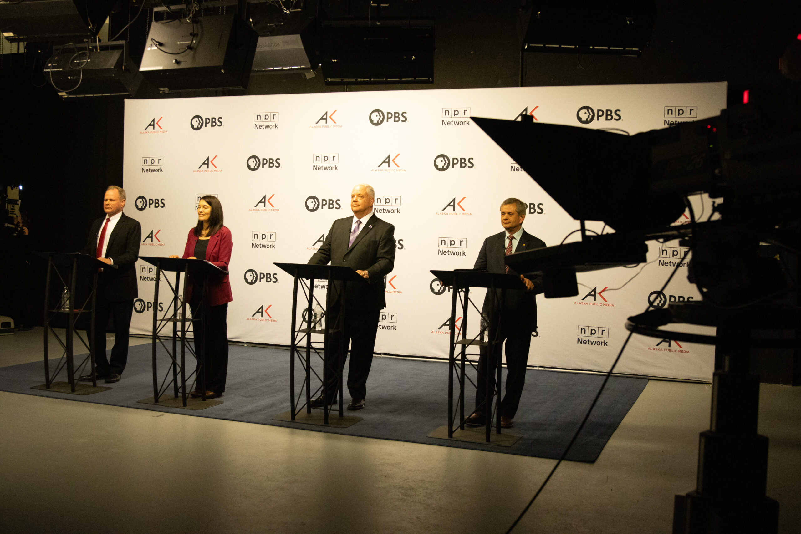 Four people, a man in a suit with red tie, a woman with a deep red blazer, a man in a suit with purple tie, and a man in a suit with a striped tie stand behind podiums in front of a white backdrop with the logos for PBS, NPR, and Alaska Public Media preparing for a debate.