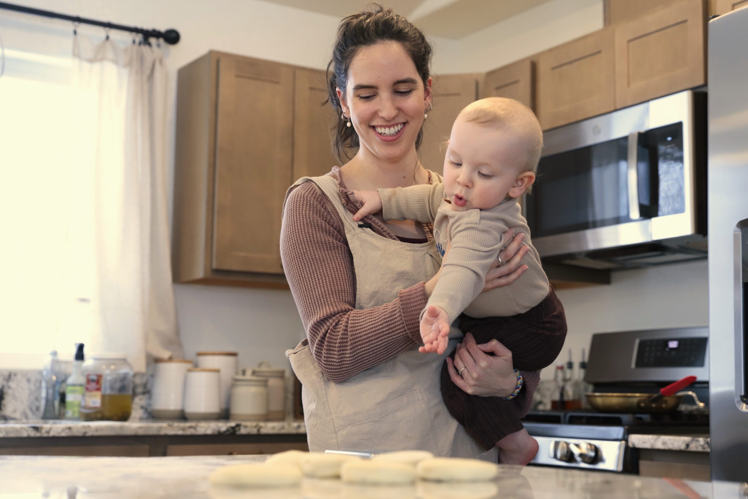 a smiling woman in an apron holds a baby reaching for doughs on a kitchen counter