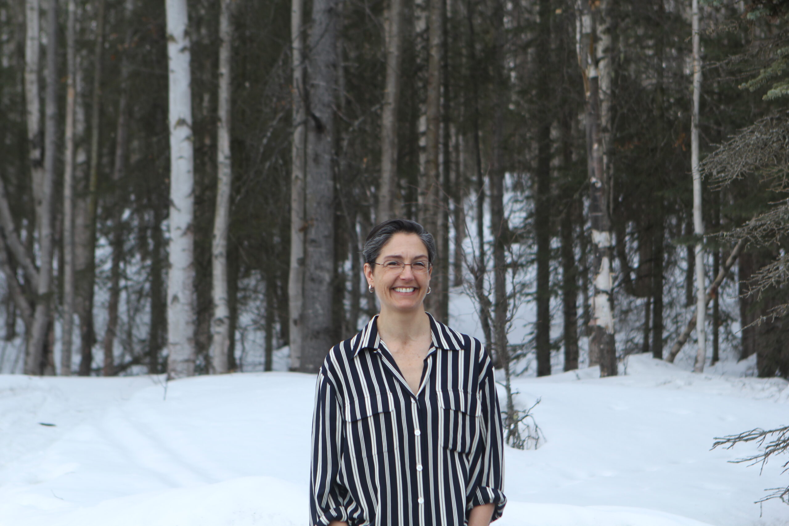 A woman in a striped shirt stands in front of snow and trees.