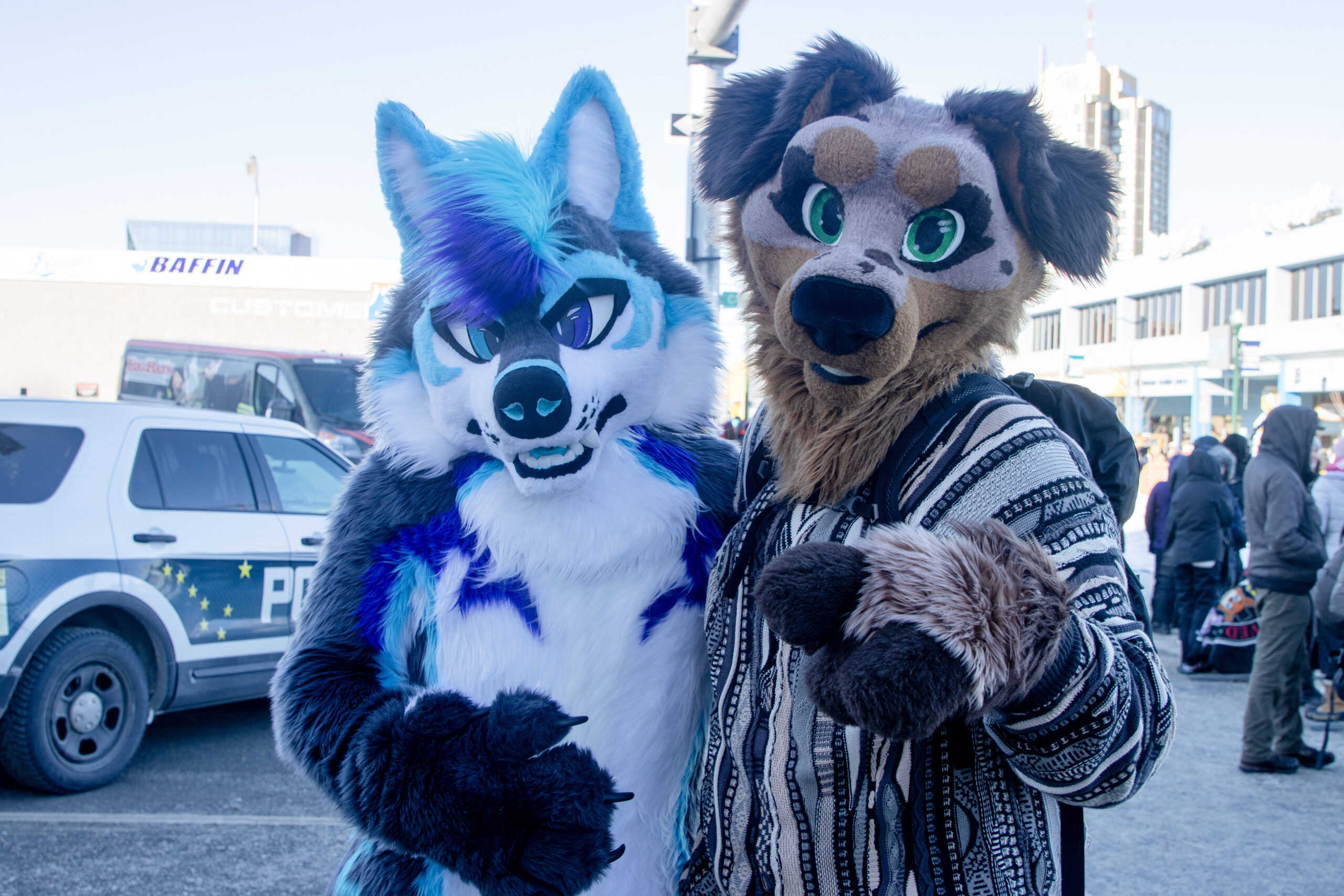 Two people dressed in wolf costumes one is blue and the other is brown pose for the camera as people walk behind them.