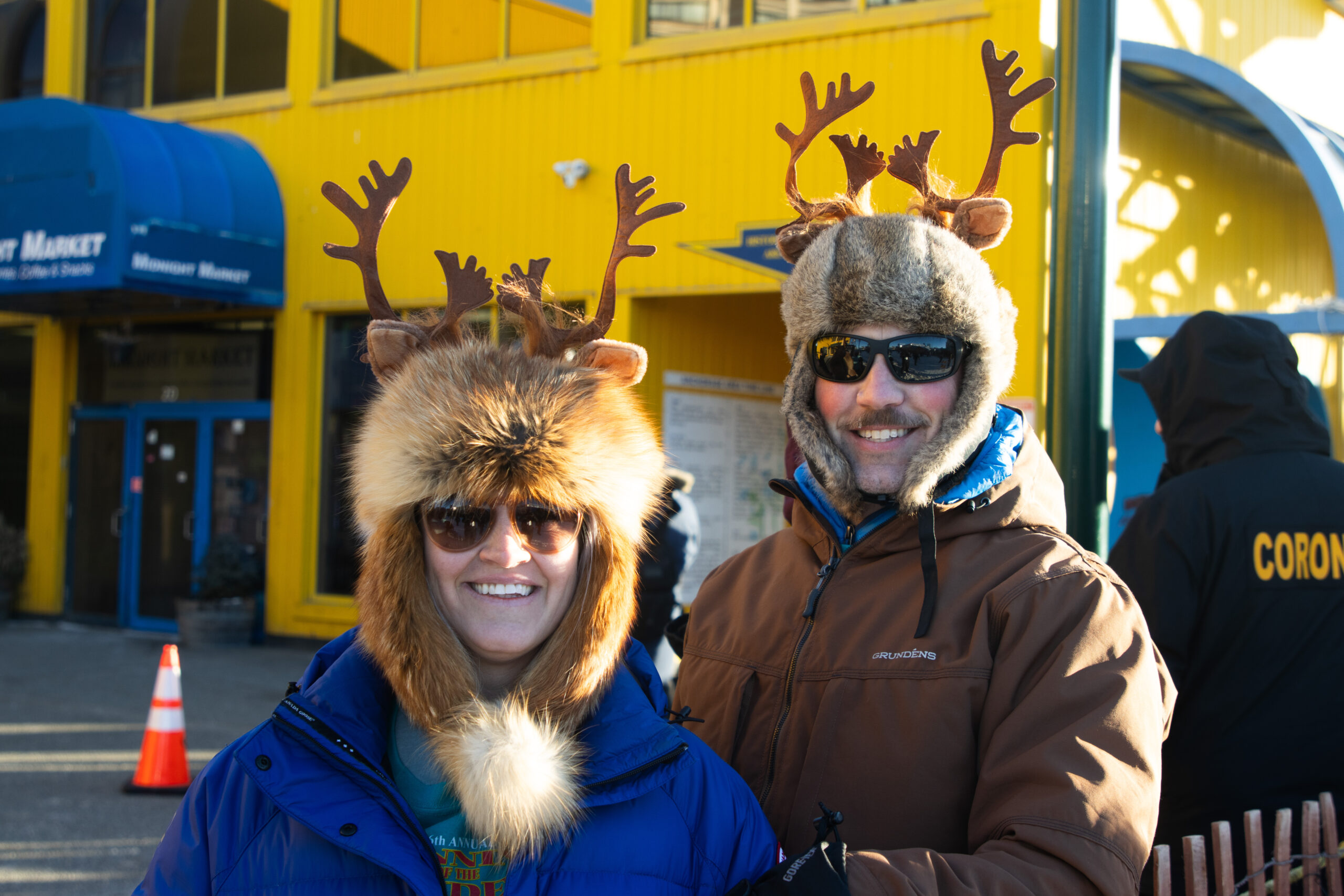 A man and a woman with fake decorative antlers pose for a photo in front of a yellow building.