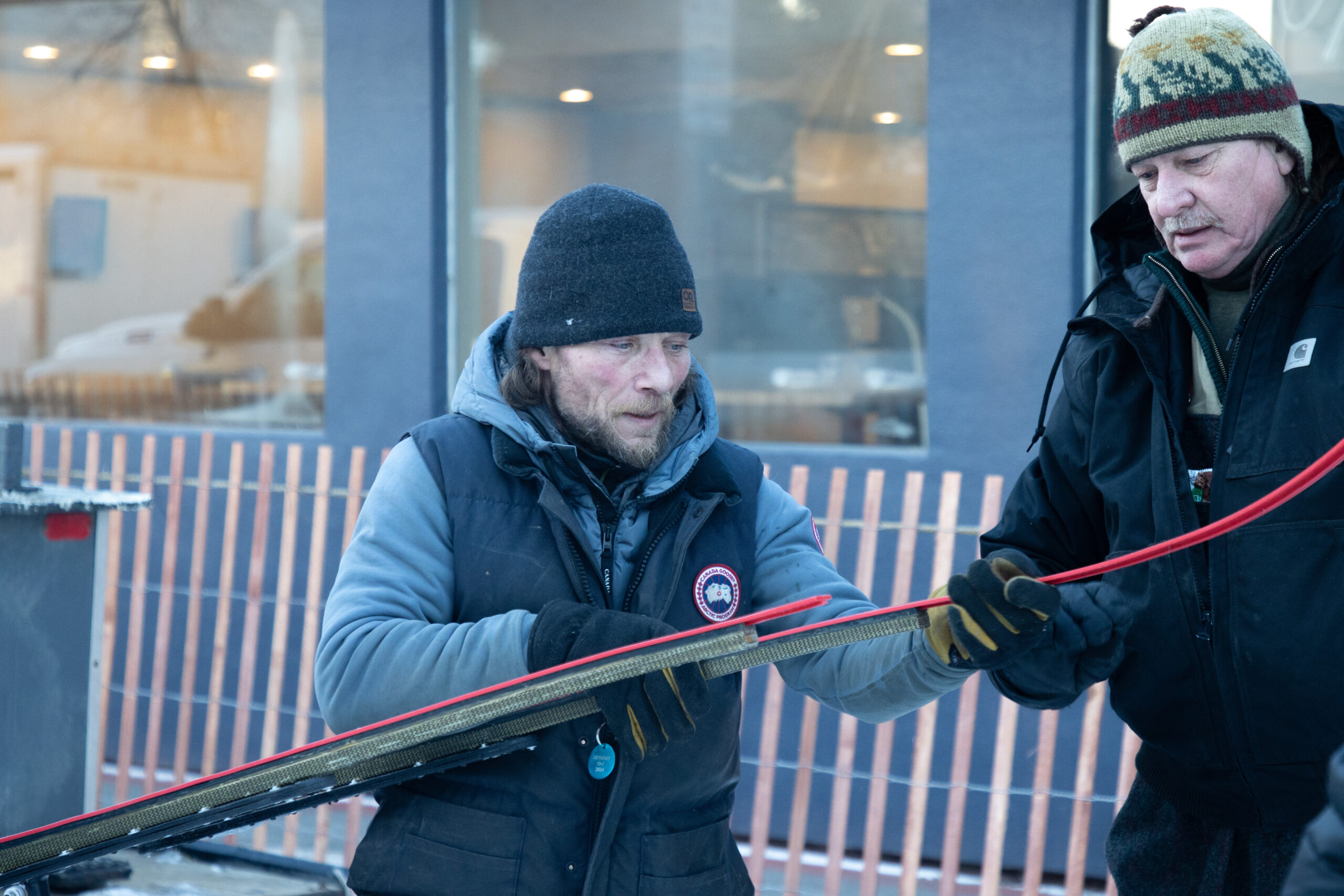 A man dressed in winter gear with a dark vest and gloves works on preparing a sled that will be used for racing.