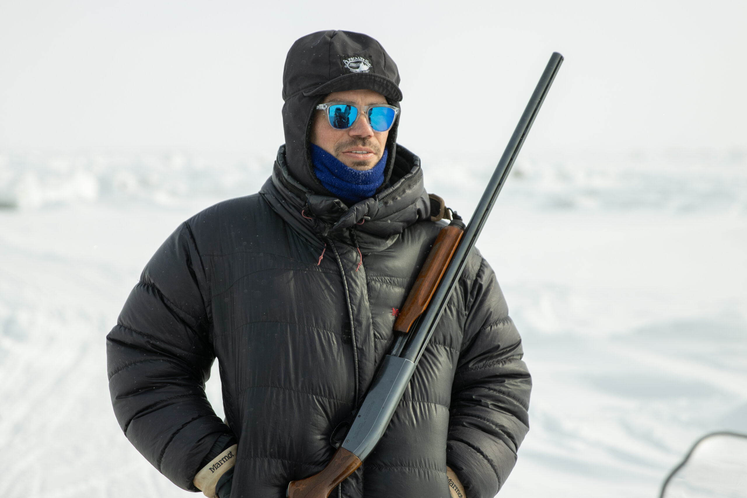 A man in a winter coat and sunglasses carries a shotgun in an arctic landscape.