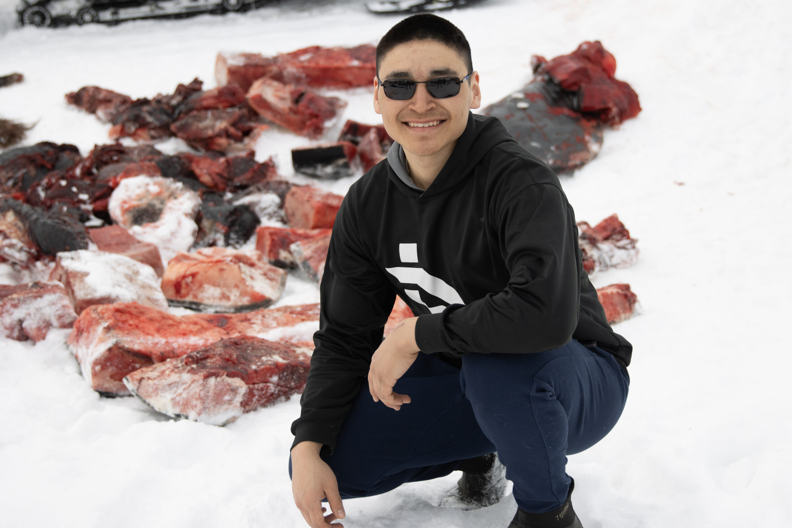 A young man in sunglasses kneels in the snow in front of pieces of red bowhead whale meat.