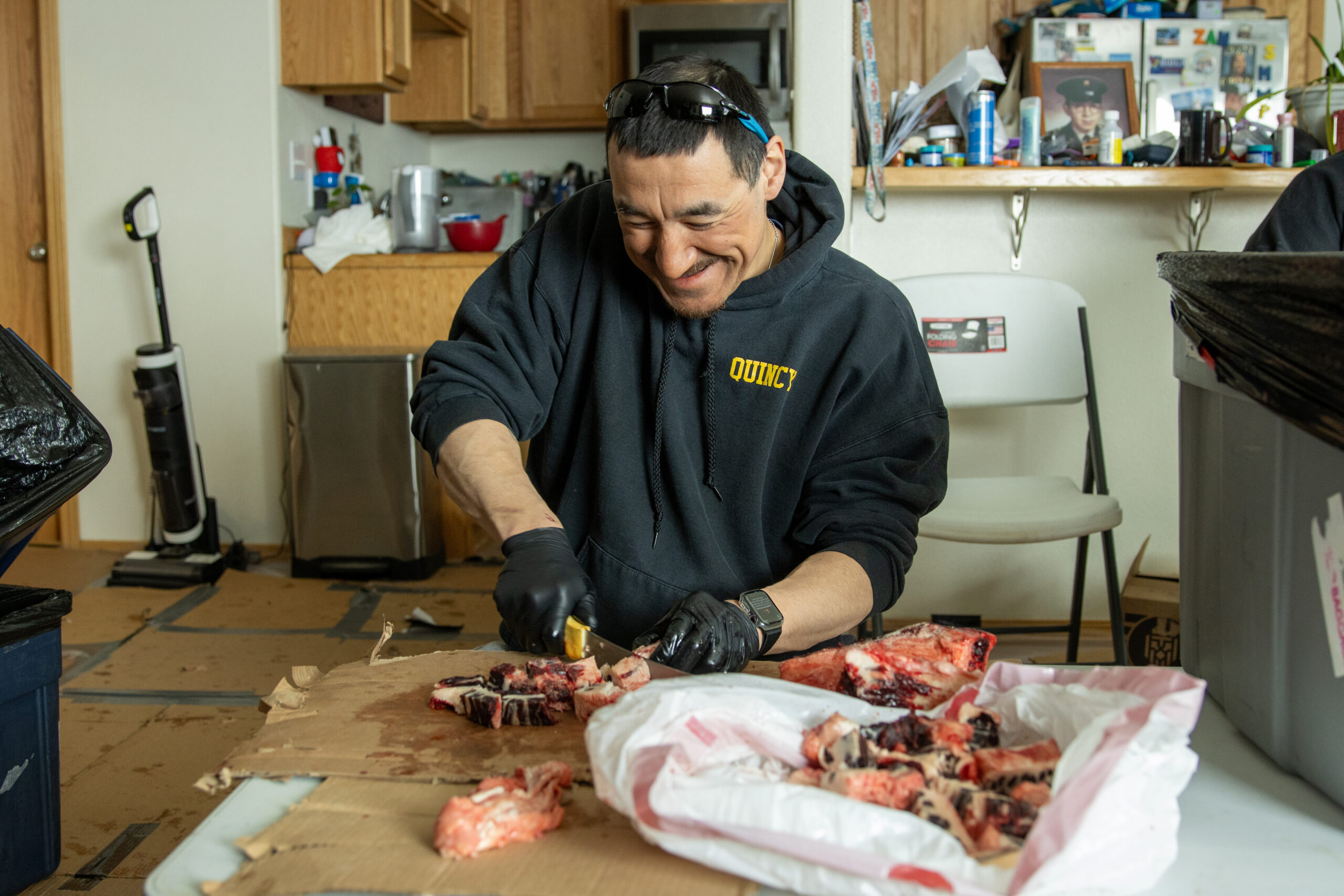 A man in a black hoodie and black rubber gloves cuts through marbled bowhead whale meat with a knife.