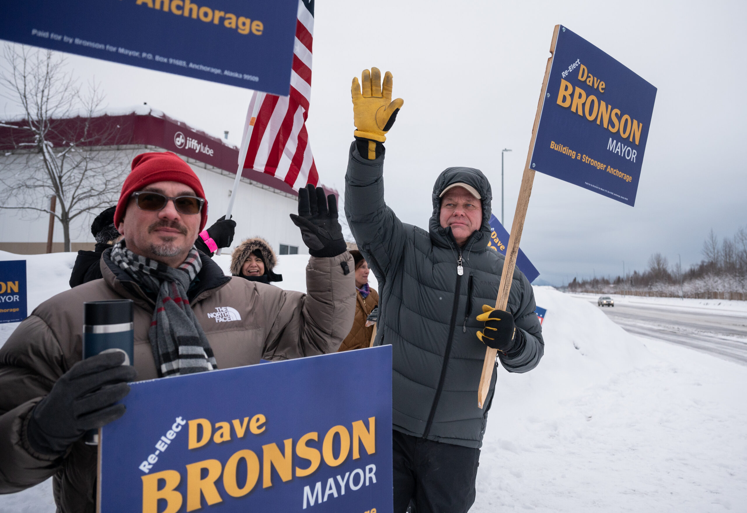 A man in a black puffy jacket stands outside waving a mayoral campaign sign