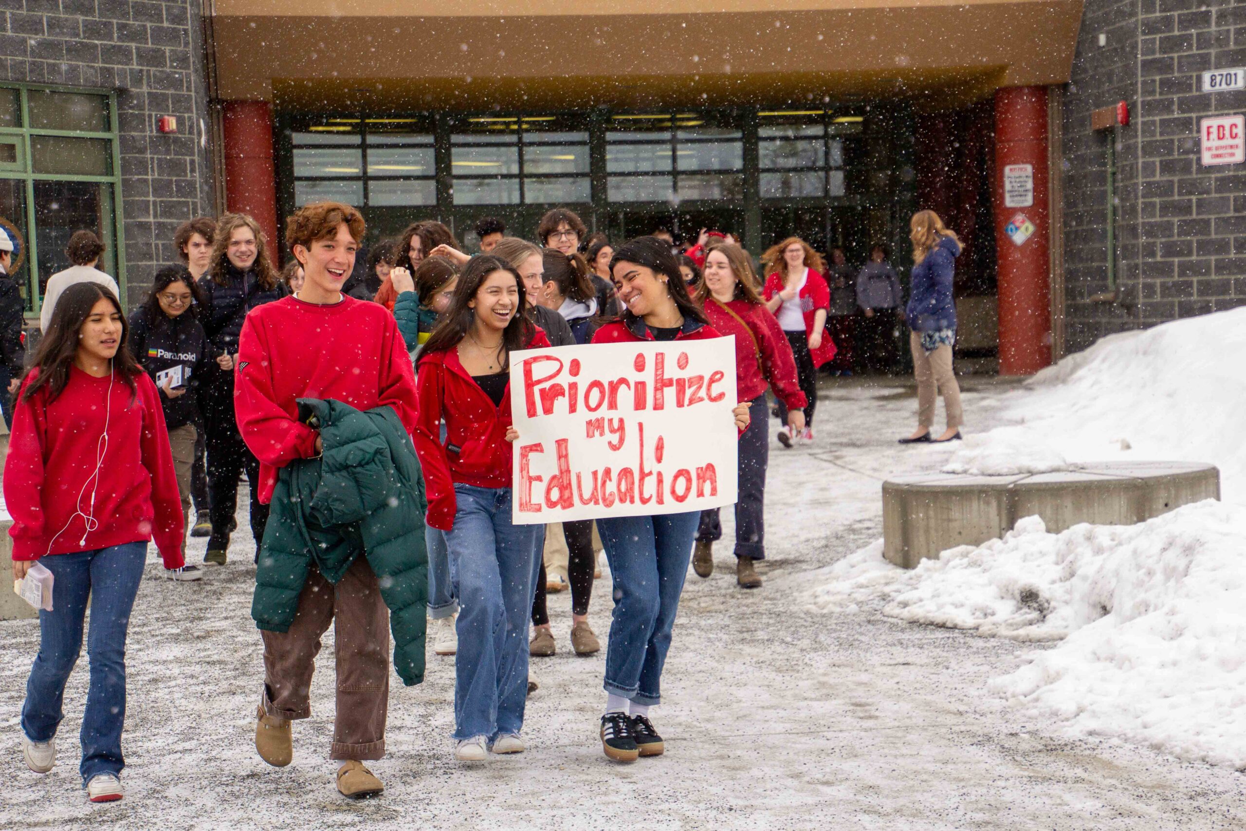 A group of students walking of their school wearing red and holding a sign that reads, "Prioritize my Education."