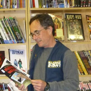 A man reads a comic in front of a shelf of comics. 