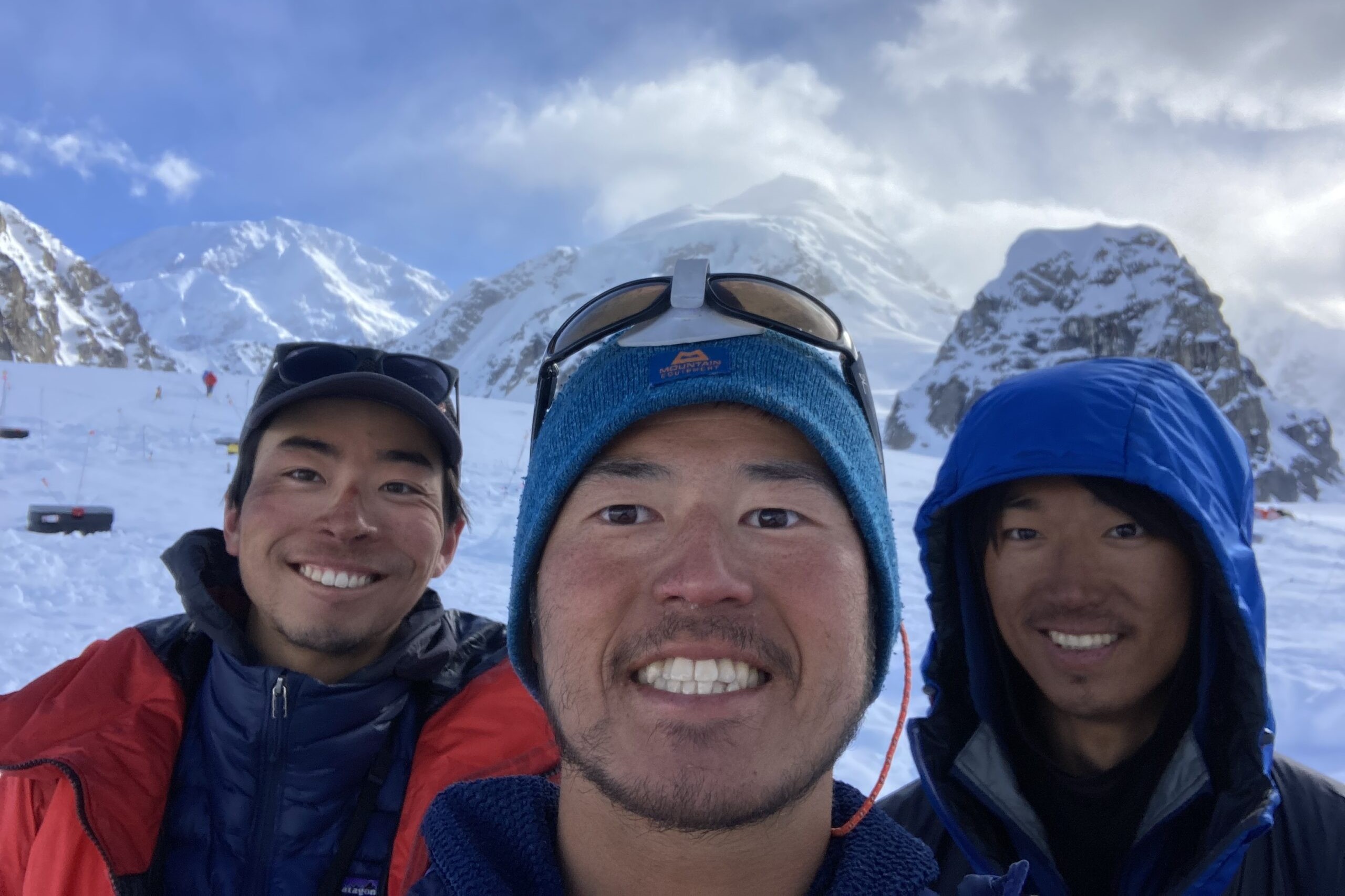 three men pose for a selfie in front of snow mountains