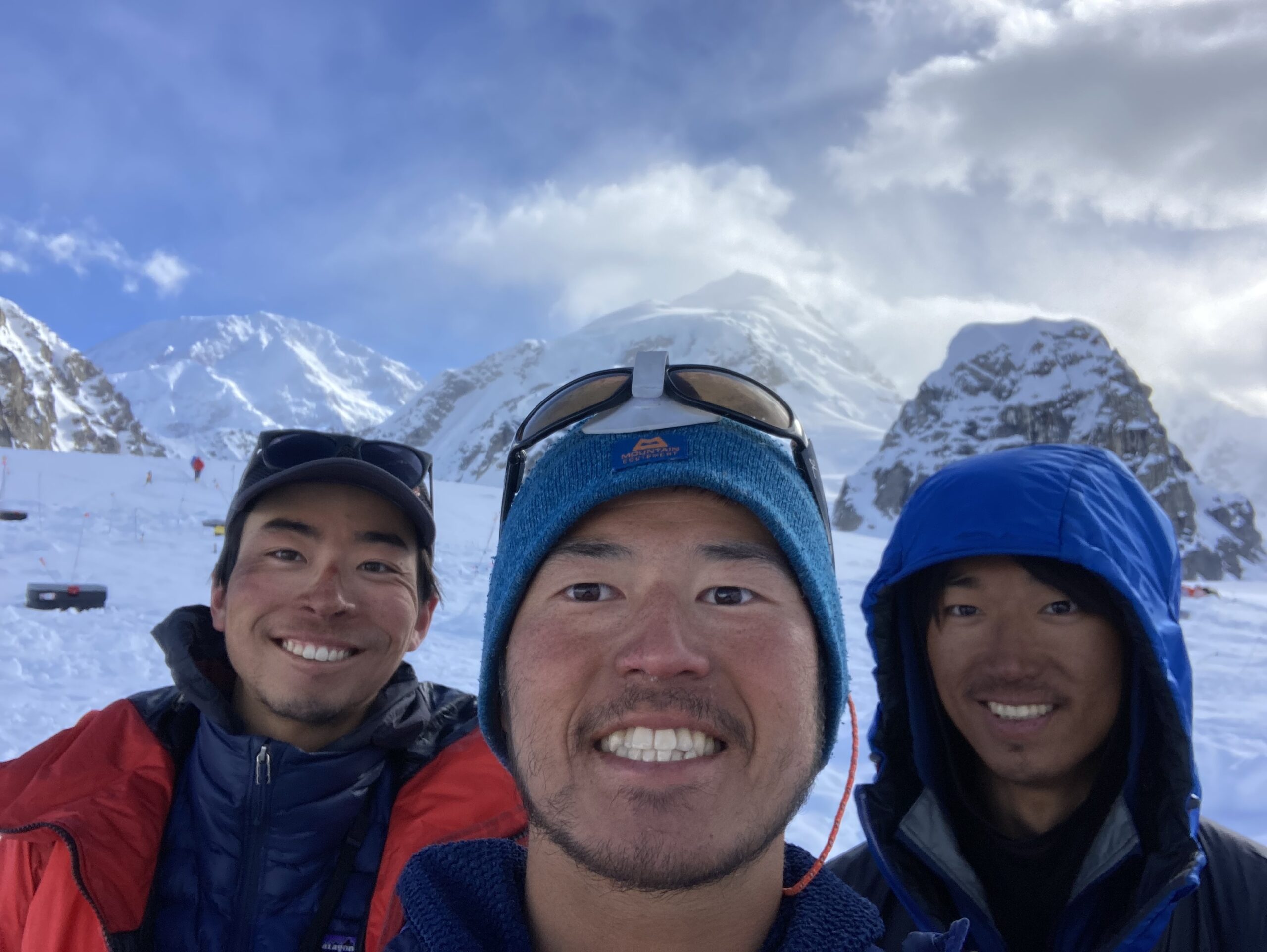 A selfie of three men wearing jackets and hats, surrounded by snow and mountains.