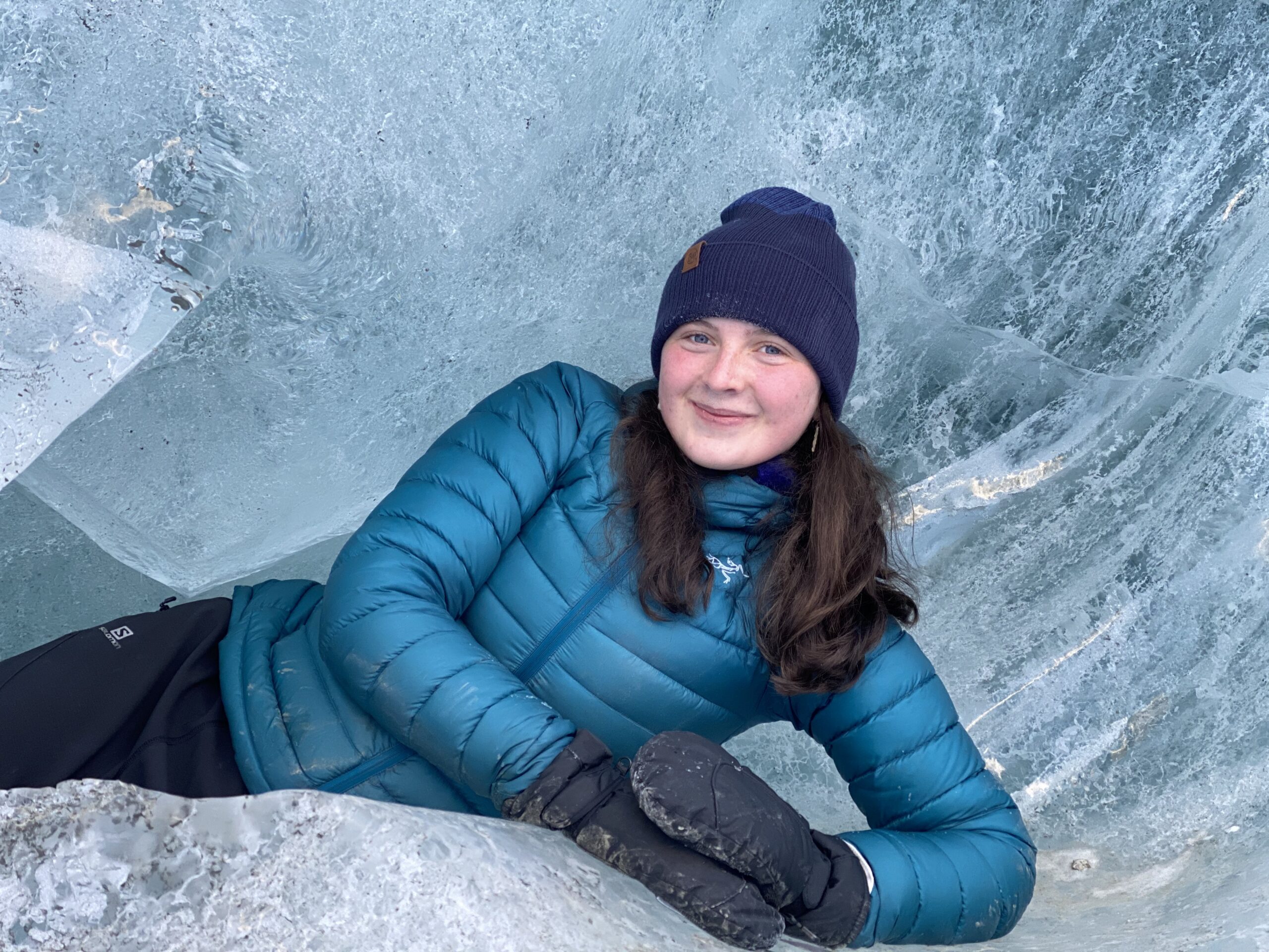 A young woman in a blue puffy jacket and navy beanie poses inside of a glacial ice cave.