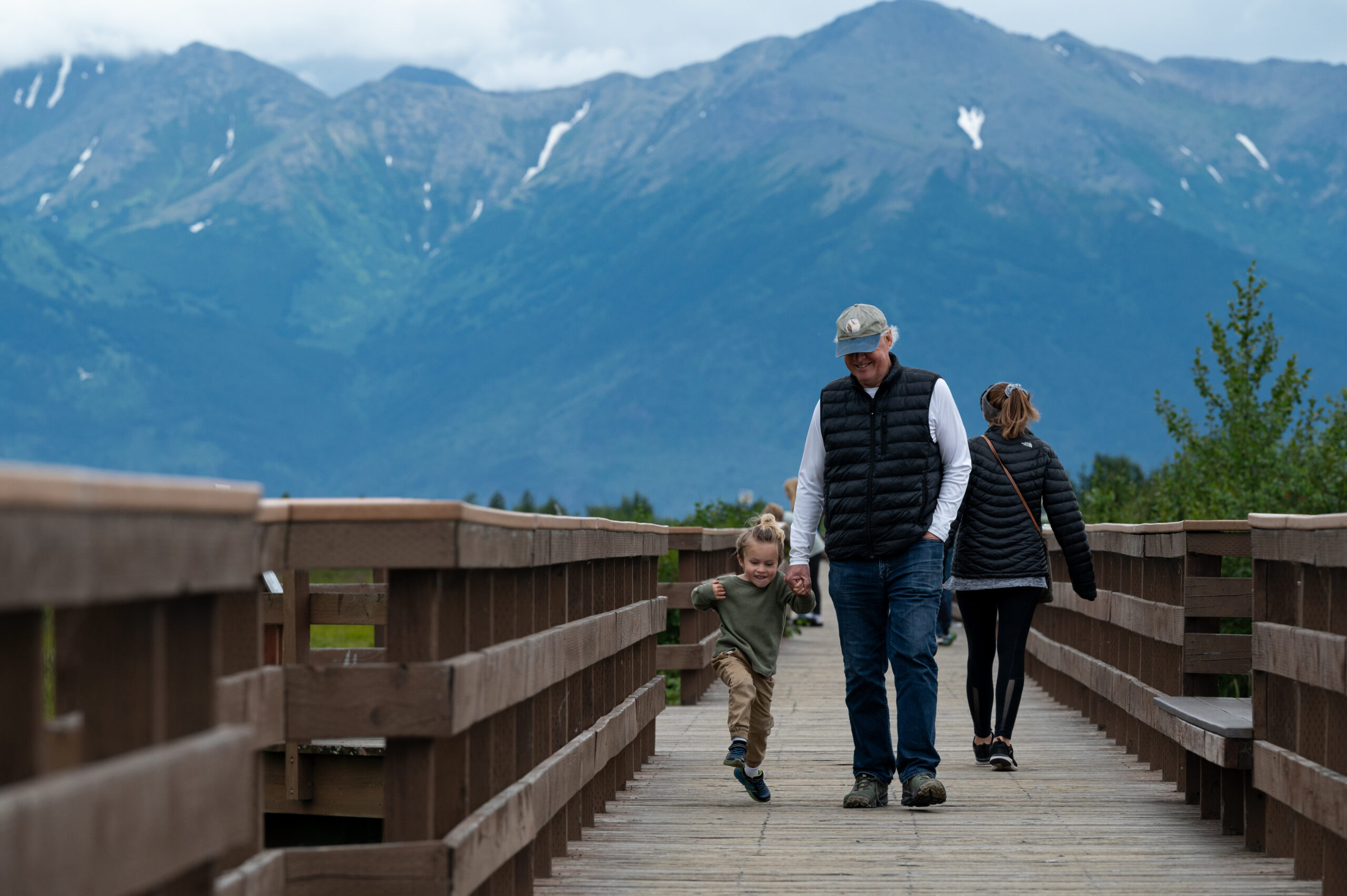 A man in a black vest walks with his grandson on a boardwalk.