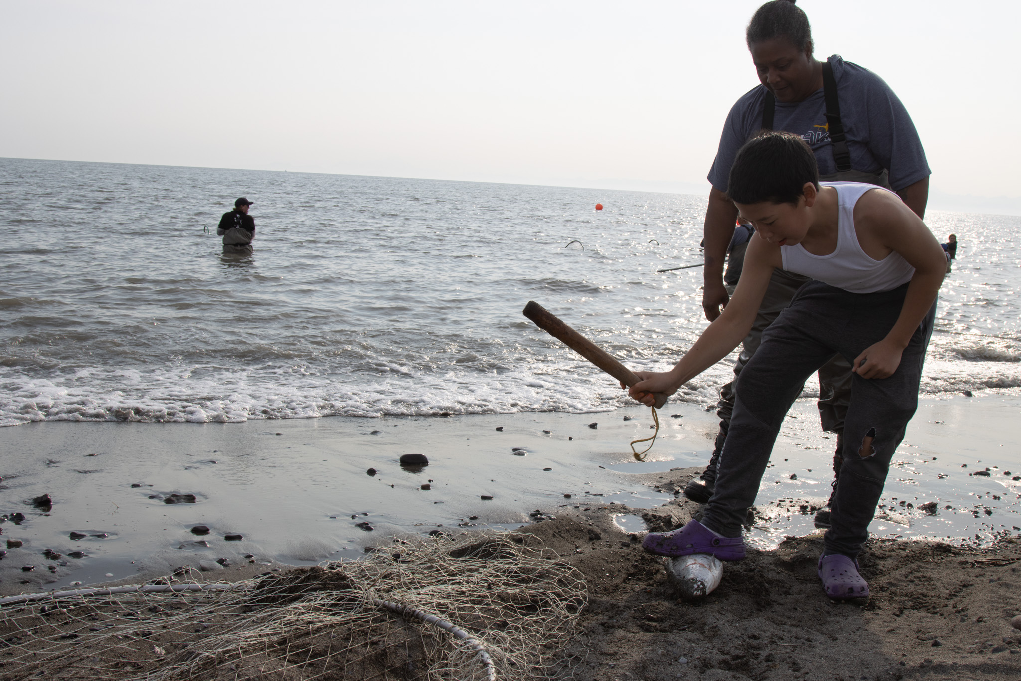 a woman stands behind a boy who is bent over with a small wooden club, about to hit a fish on the sand