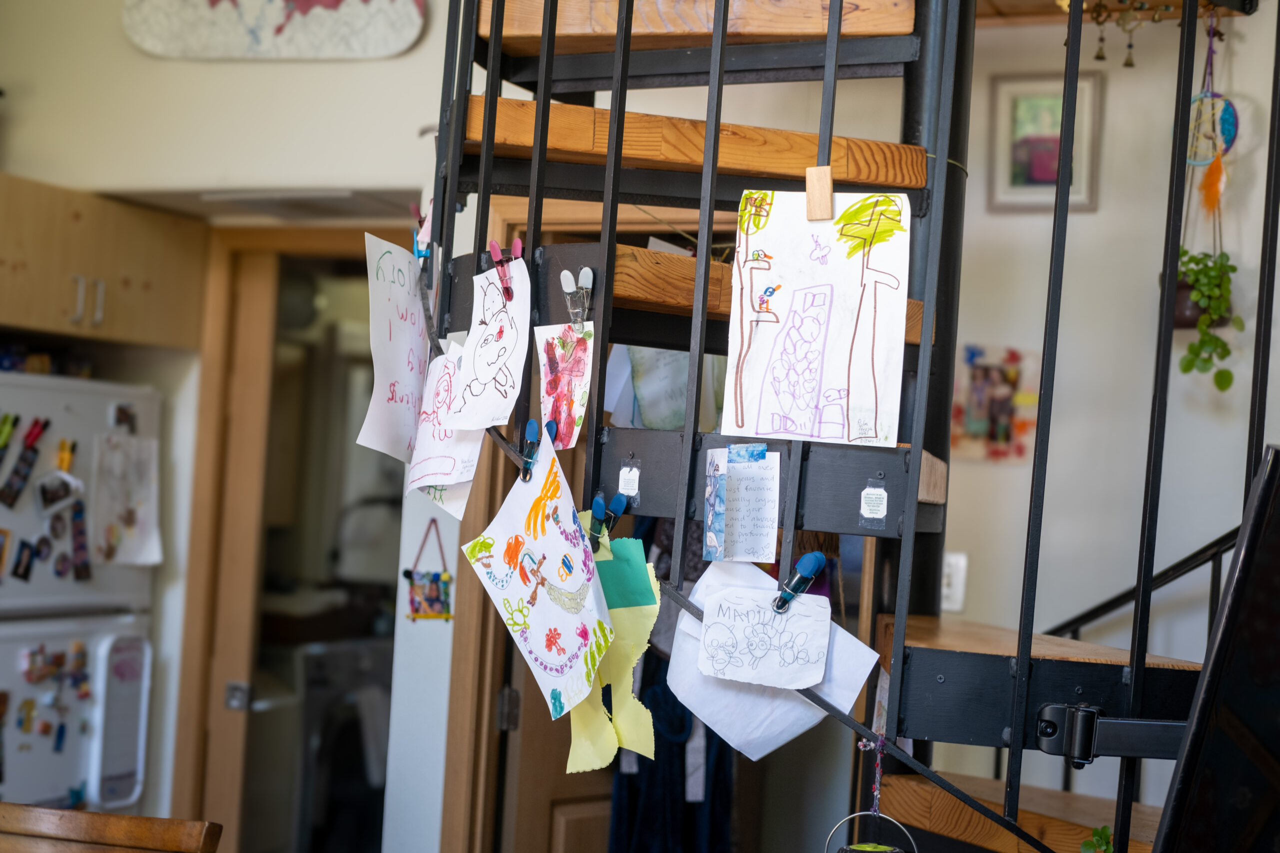 Several colorful children's drawings are pinned to a spiral staircase inside a home. A doorway and fridge with lots of pictures on it are in the background.