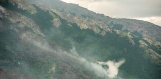 A yellow and red super scooper plane, dumping water over smoke emerging from mountainous terrain