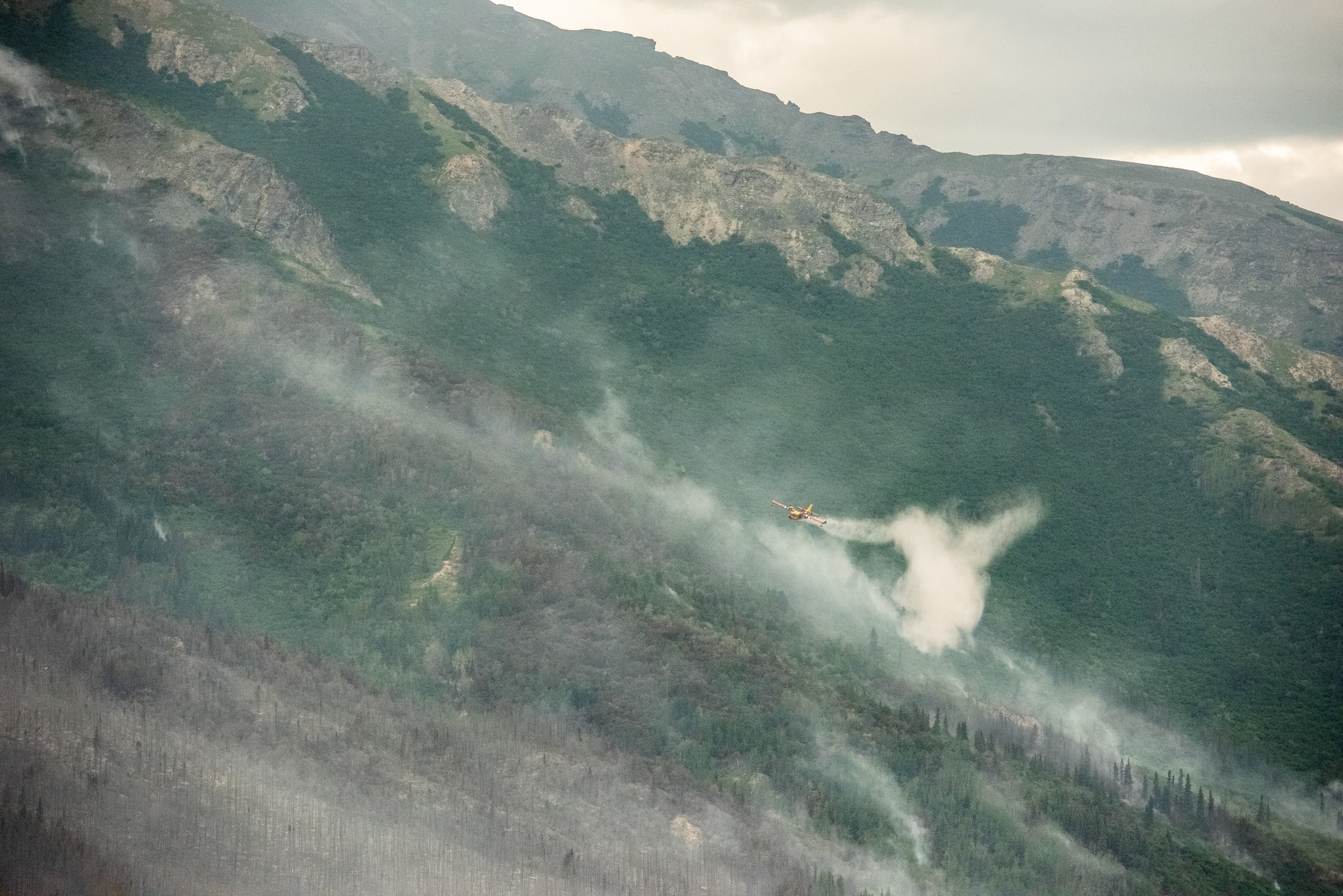 A yellow and red super scooper plane, dumping water over smoke emerging from mountainous terrain