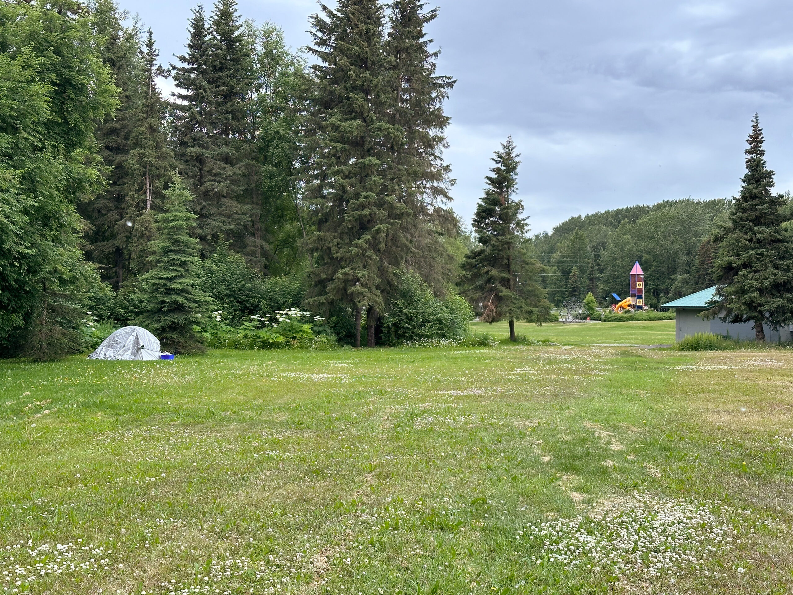 A tent with a gray tarp over over it set up with a kids playground in the distance.