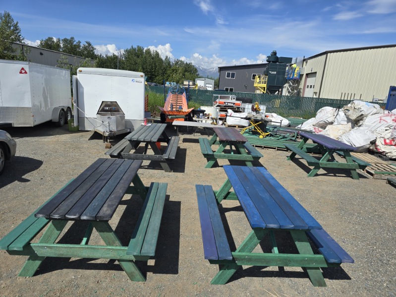 Five picnic tables are outside in a gravel lot. The picnic tables are made of plastic lumber, which is various different shades of blue and green.