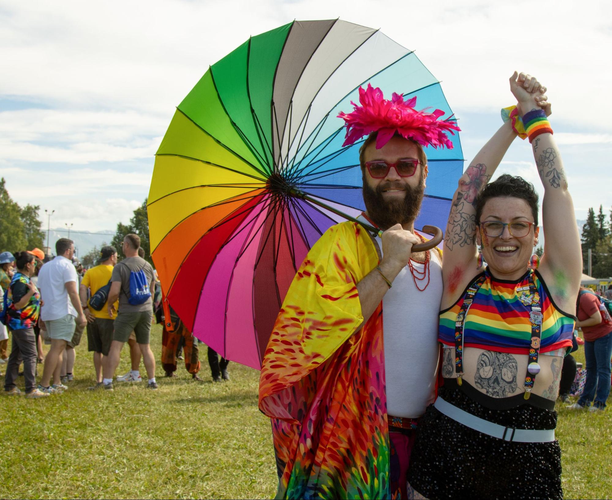 Two adults wearing rainbow clothing and one holding an umbrella smile in a photo.