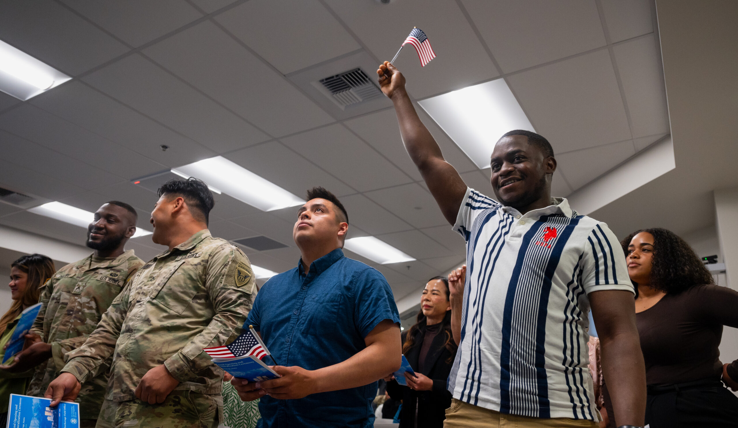 Four men stand in an fluorescently lit office room. the man on the right is waving an American flag with a big smile on his face. two other men are wearing US Military uniforms.