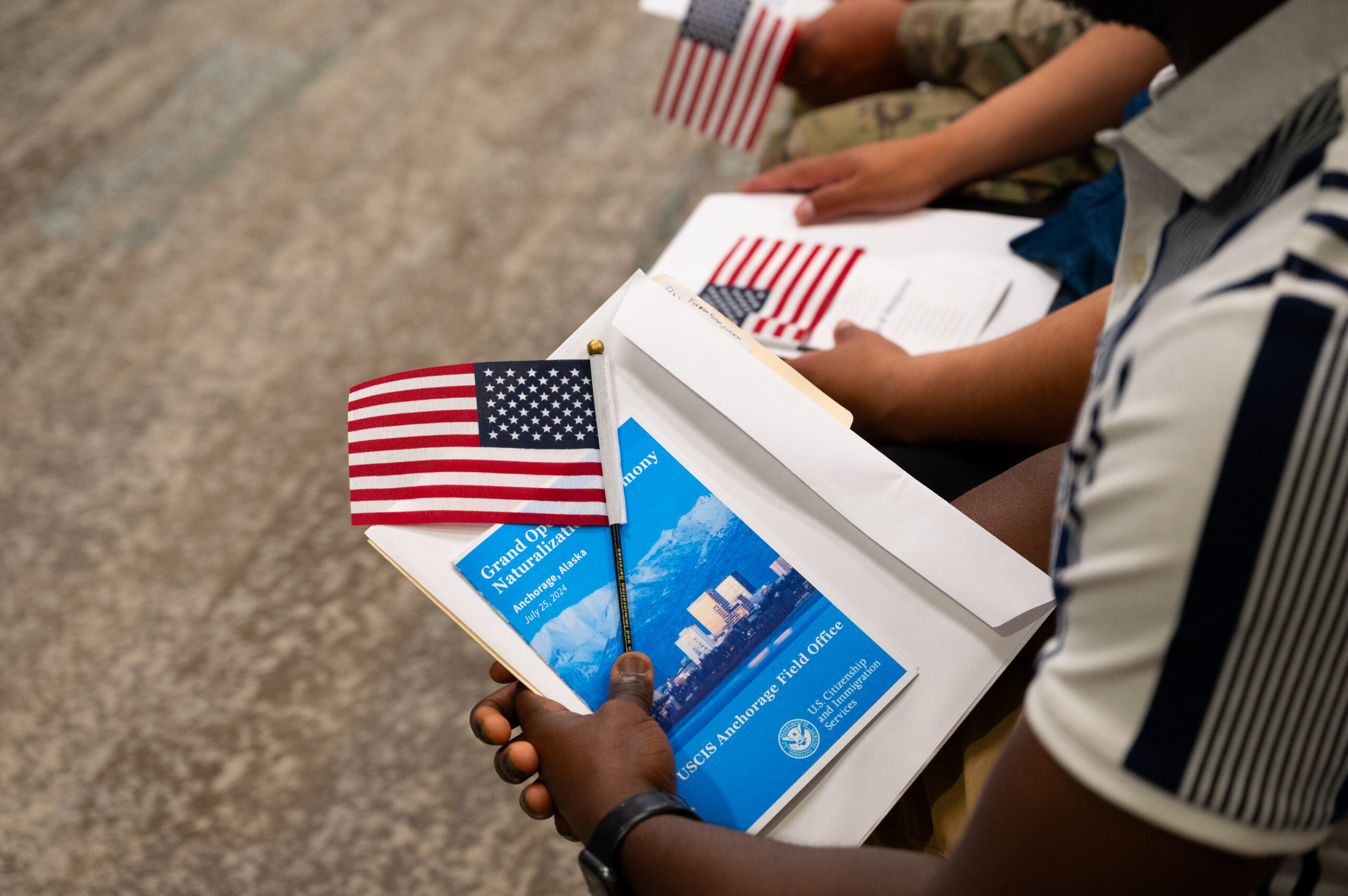 A close-up on some hands holding a large white envelope with a pamphlet on top of it that says "Naturalization Ceremony USCIS Anchorage Field Office," and on top of that a small American flag on a stick