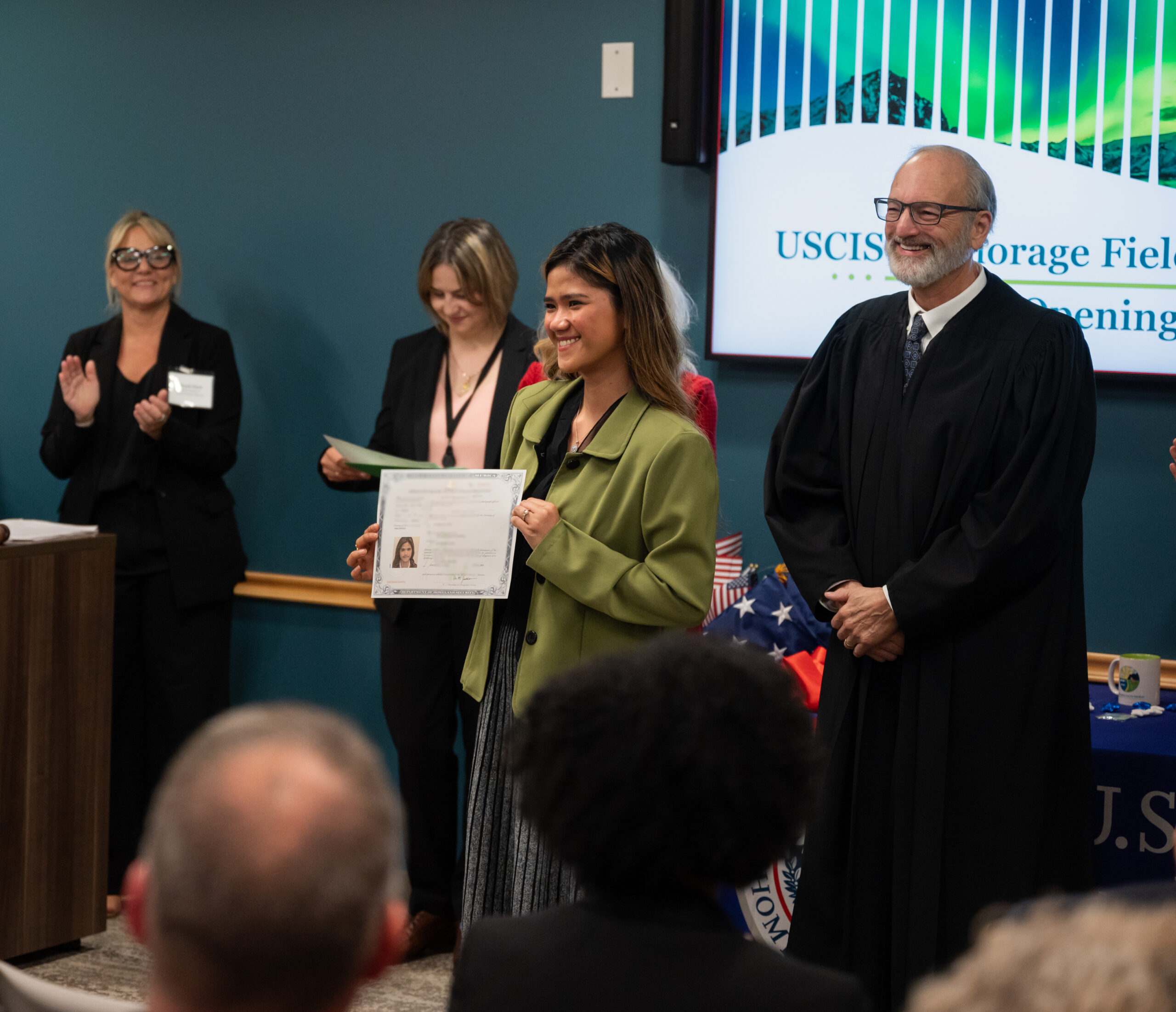 a female successful a greenish overgarment smiles while holding a certificate of naturalization. Behind her is nan judge who officiated her citizenship and 2 women who are clapping for her.