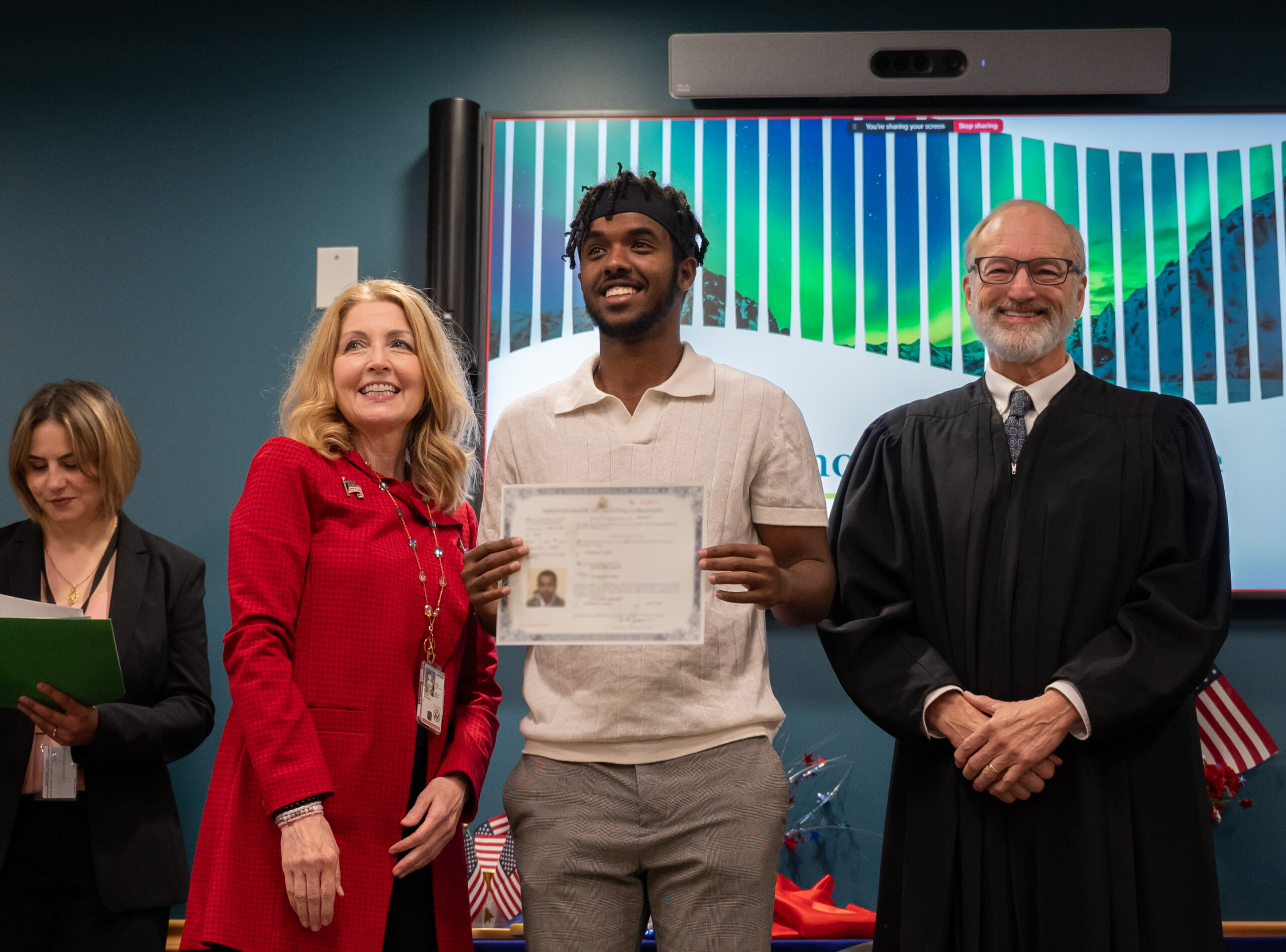 a man stands holding his certificate of naturalization. connected his correct of him is nan judge who officiated his citizenship and connected nan near is simply a female successful a reddish coat. each 3 are smiling and posing