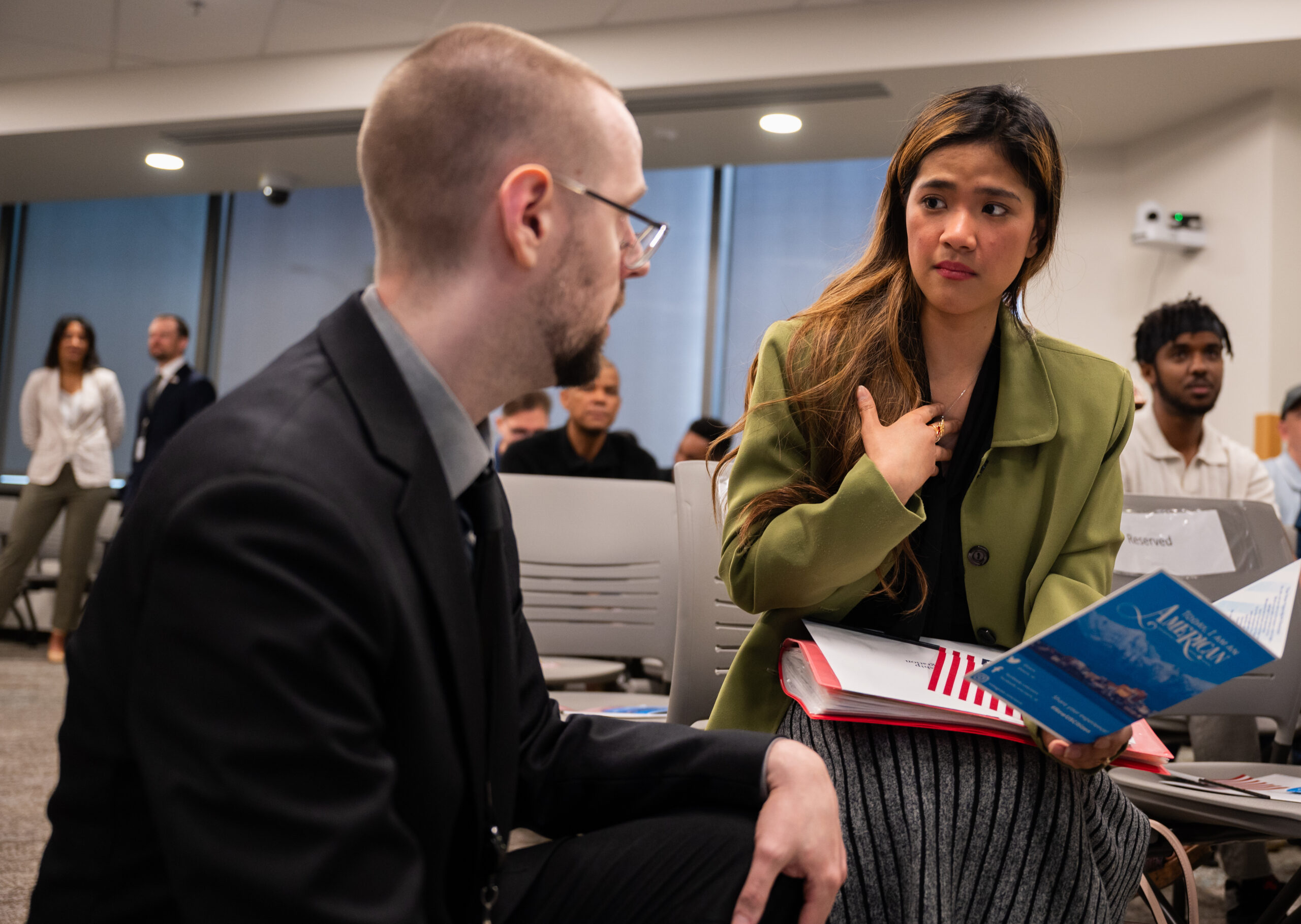 a woman in a green jacket listens with a serious expression as a man with glasses squats down to tell her something.